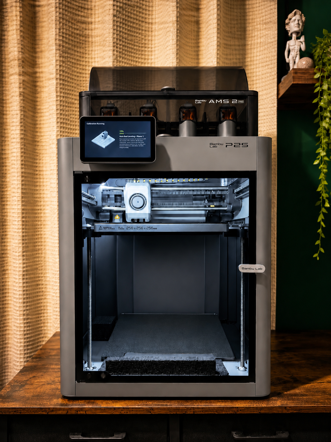 A black and gray 3D printer with a touchscreen, on a wooden table, in front of beige curtains and a green wall with a shelf holding a plant and a figurine.