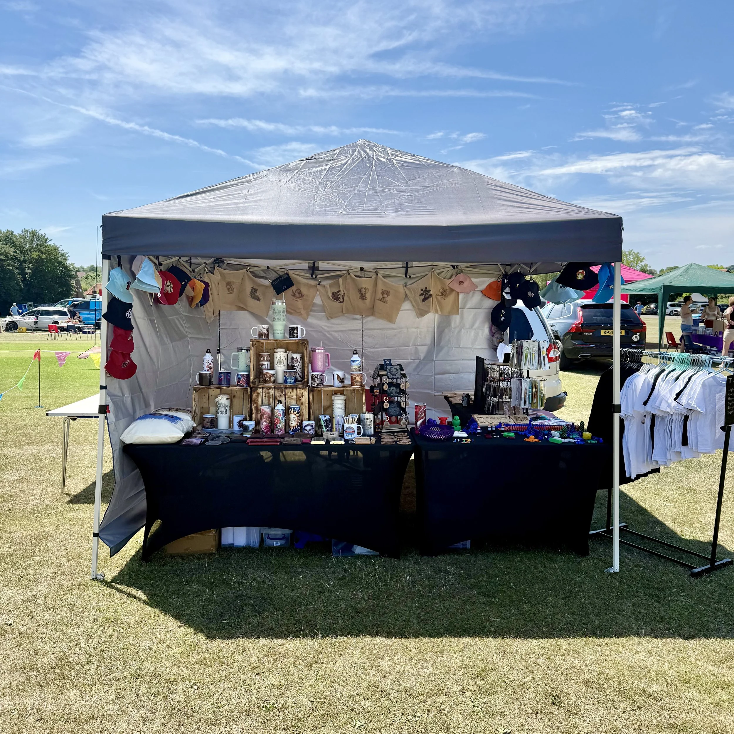A pop-up market stall with a gray canopy selling various items including hats, mugs, and decorative objects, set in a grassy outdoor area on a clear, sunny day.