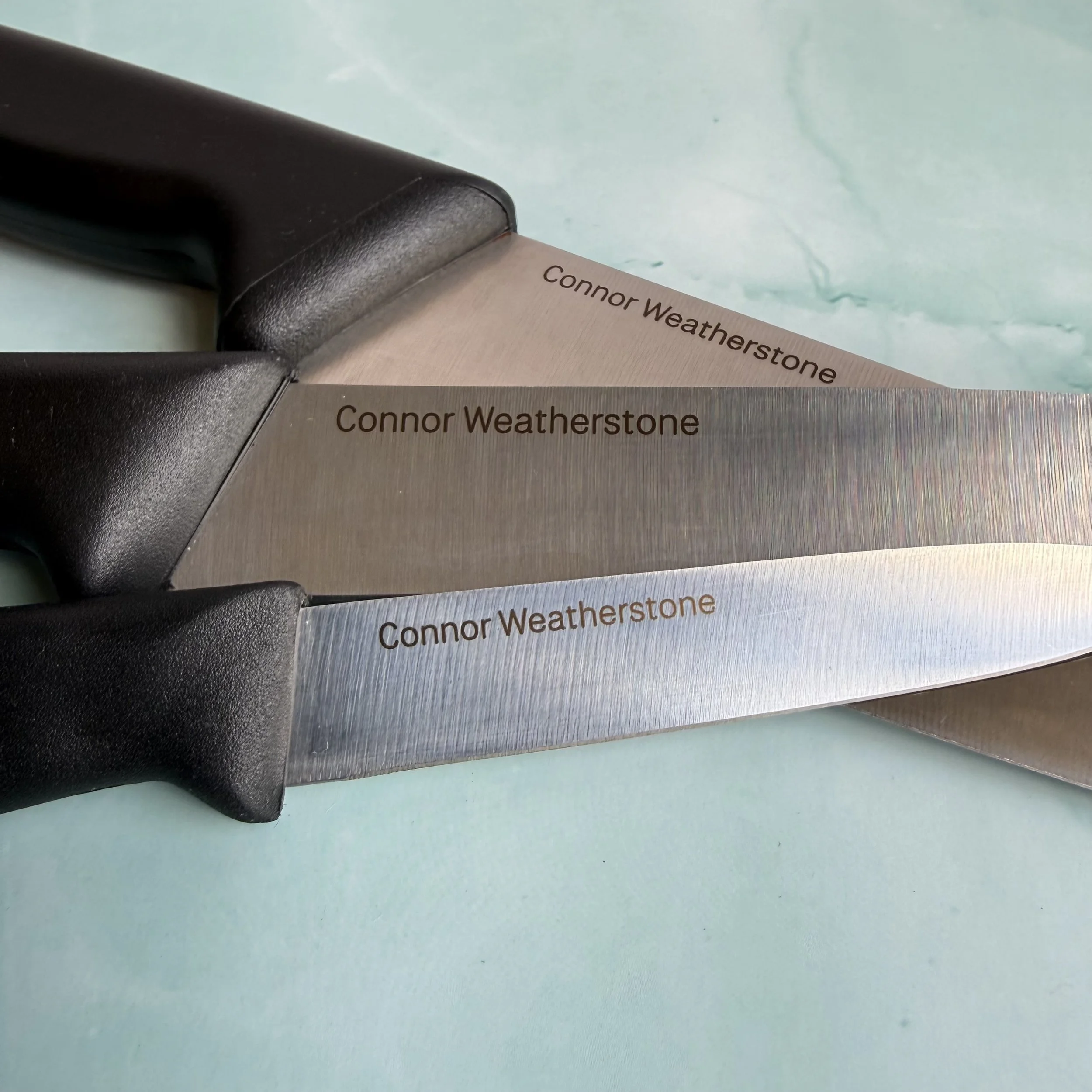 Close-up of a stainless steel kitchen knife with the name "Connor Weatherstone" engraved on the blade, lying on a light blue surface.