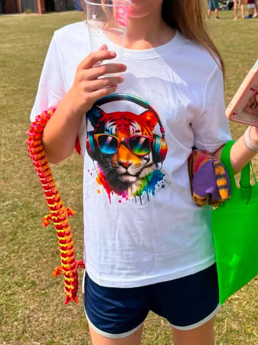 Person wearing a white T-shirt with a graphic of a tiger wearing sunglasses and headphones, holding a clear plastic cup, with a colorful lei and holding a phone in one hand and a bright green bag.