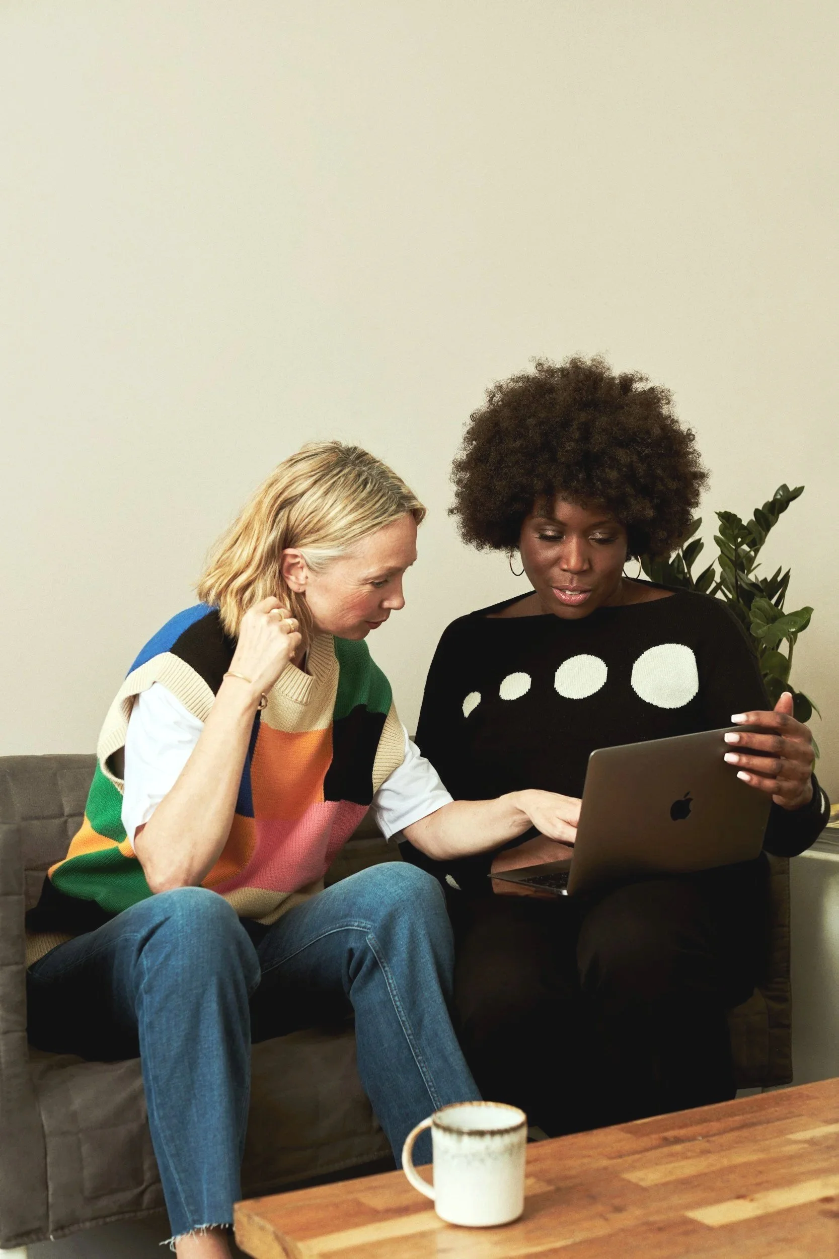 Two women are sitting together on a couch, looking at a laptop. One woman has blonde hair and is wearing a colorful striped sweater, while the other woman has curly hair and is wearing a black sweater with white dots. There is a plant in the background and a coffee mug on the table in front of them.