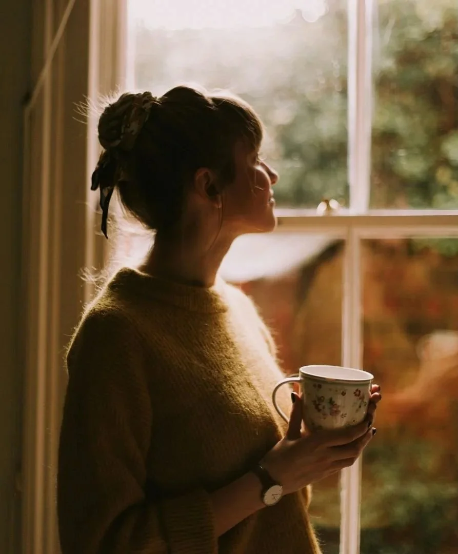 A woman sitting quietly with a warm cup of tea, gazing out a window in a moment of reflection.