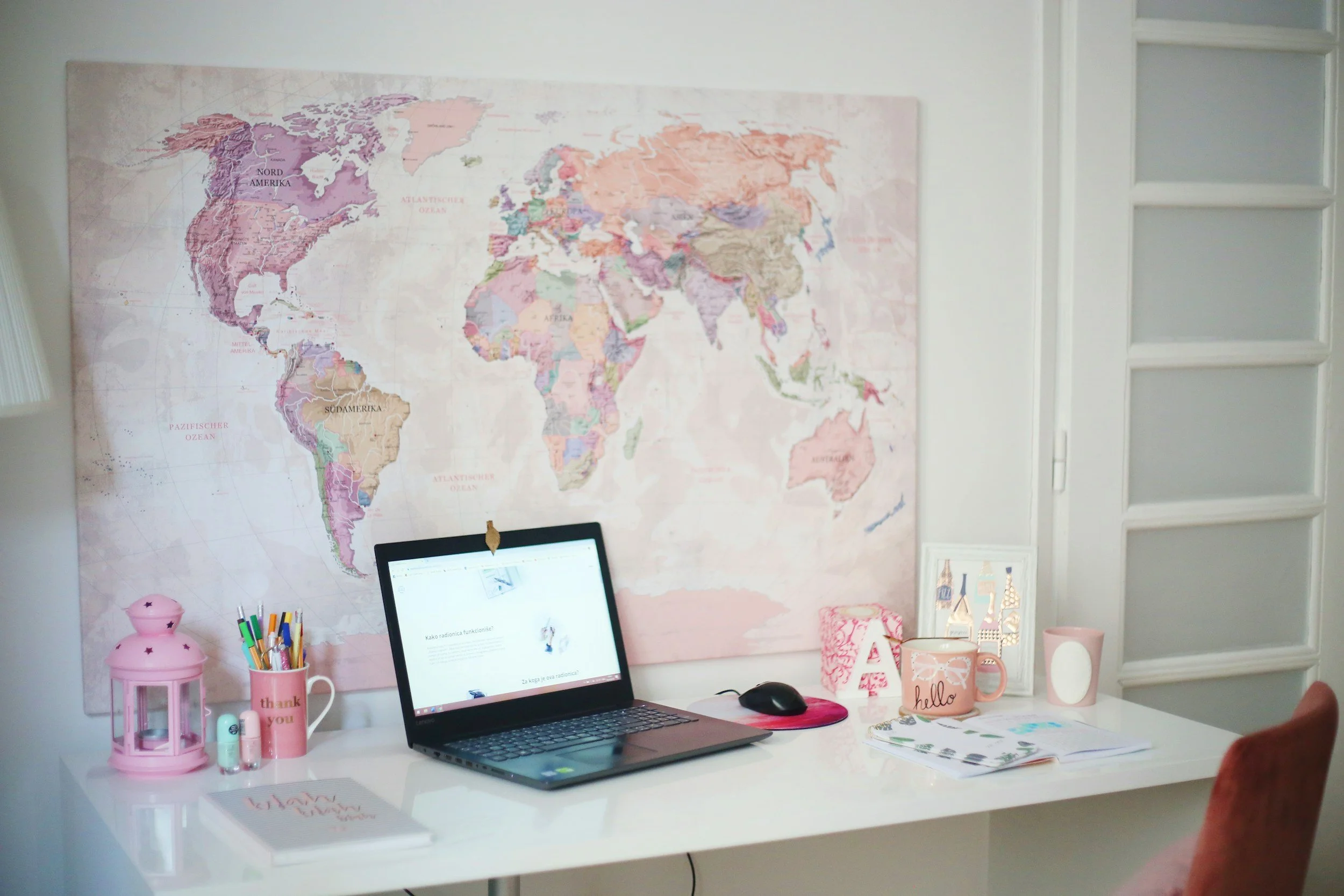 A white desk with a laptop, colorful pens in a pink holder, a pink lantern, and various cups. Behind the desk, a large world map is mounted on the wall with pastel colors. To the right, white built-in shelves are partially visible, and there are framed decor items and a notebook on the desk.
