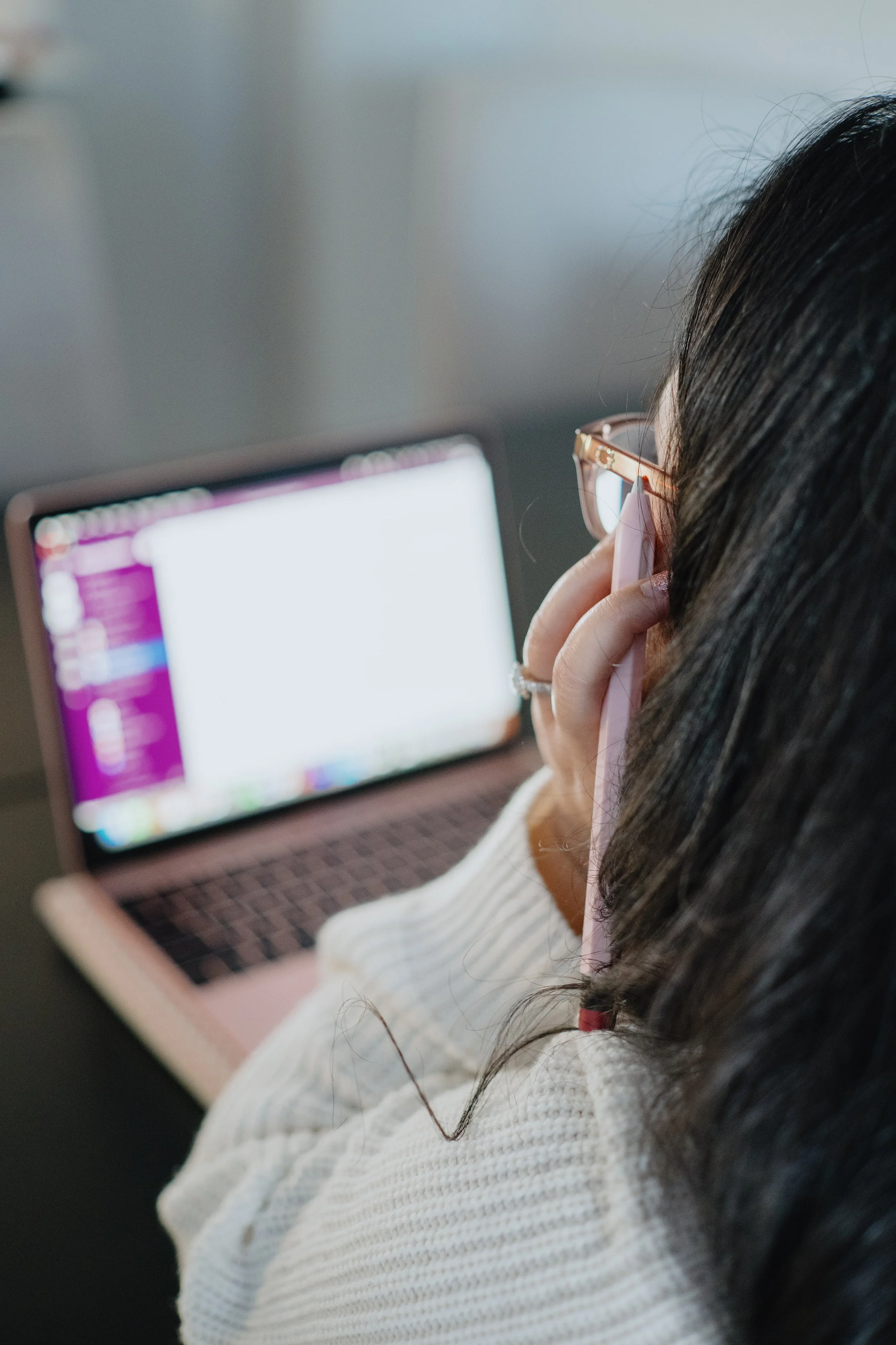 A woman with dark hair and glasses, wearing a white sweater, is resting her chin on her hand while looking at a laptop screen. The laptop screen displays a colorful interface.