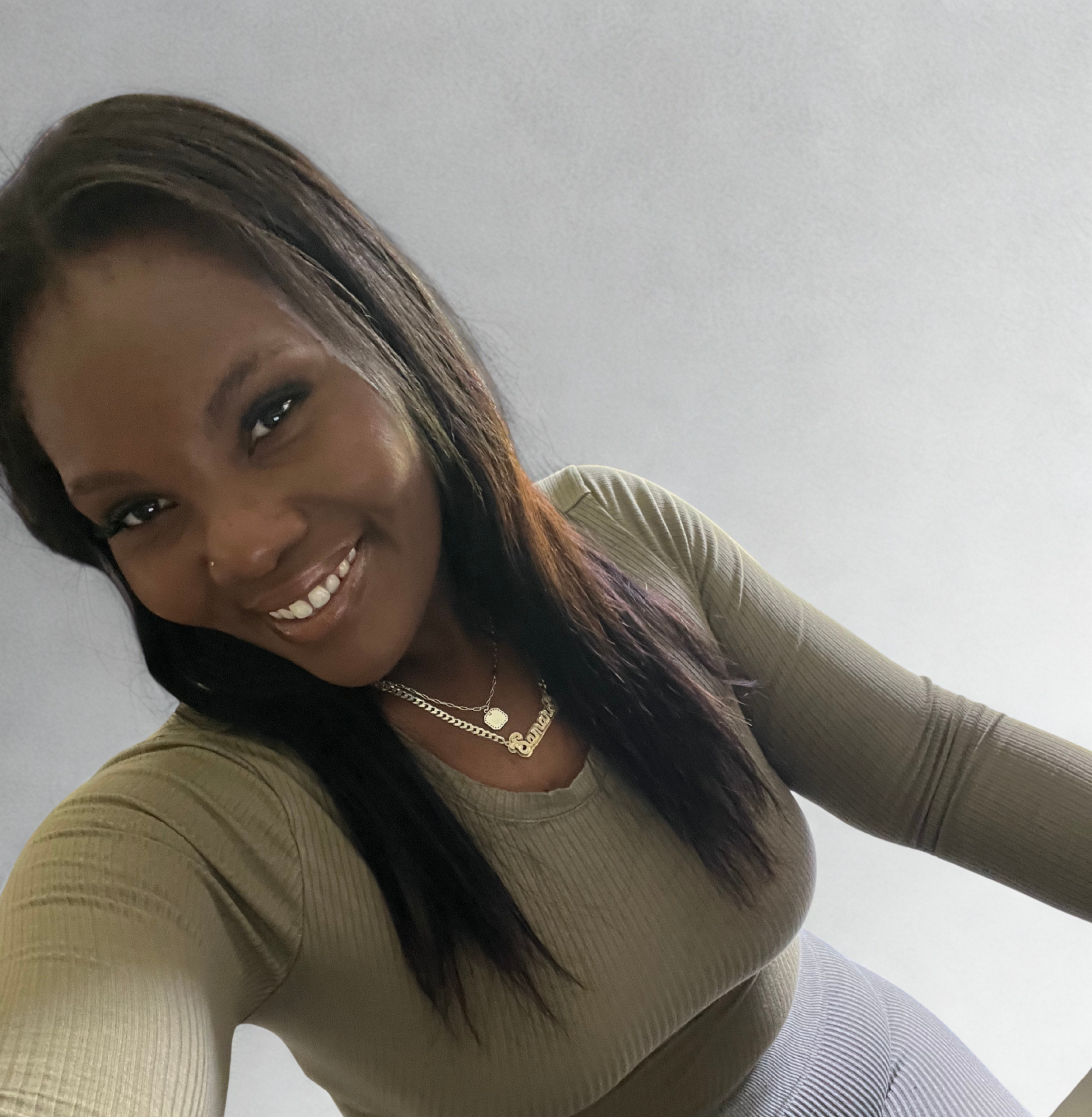 A woman smiling with long black hair and wearing a tan ribbed long-sleeve top, accessorized with layered necklaces, standing against a plain white wall.