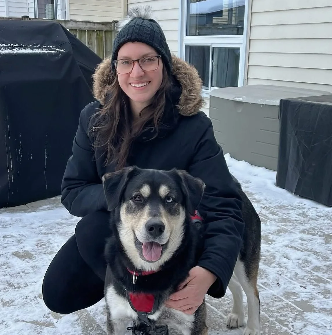 A smiling woman wearing a black winter coat, glasses, and a knit hat with a pom-pom, kneeling outdoors on snow, holding a black and white dog with heterochromatic eyes and a red harness.