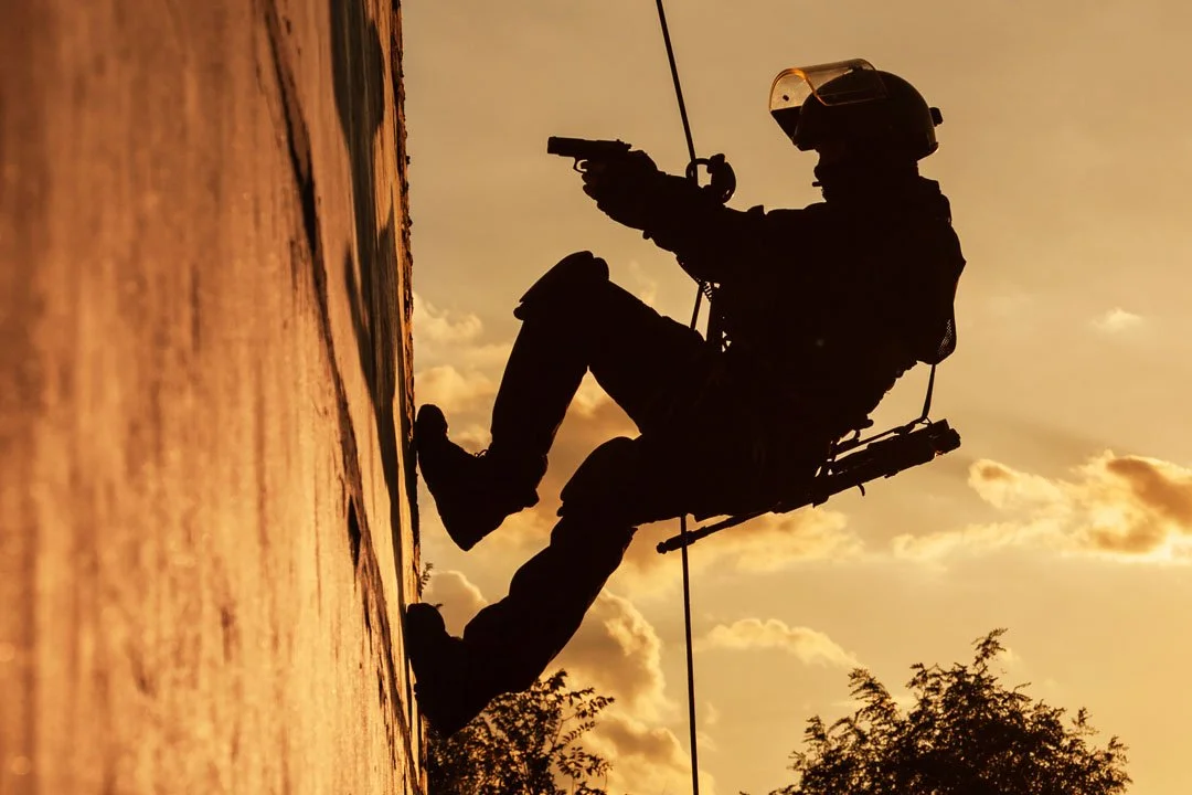 Silhouette of a soldier wearing tactical gear and a helmet, sitting on a rope and aiming a handgun during sunset.
