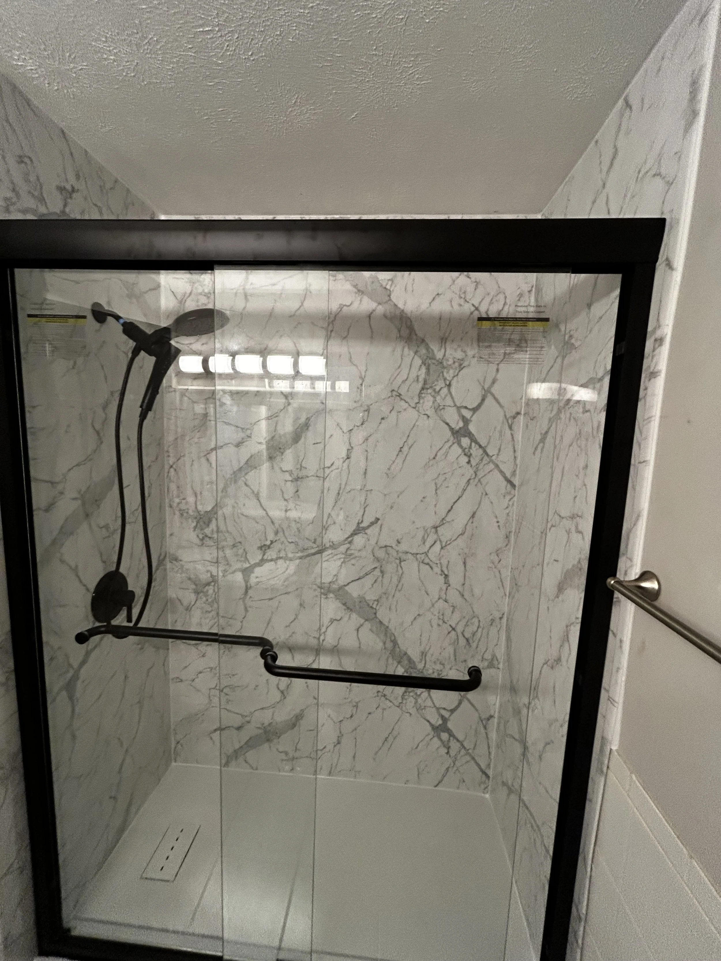 A marble-finished shower with a glass door, a black grab bar, and a handheld showerhead mounted on the wall.
