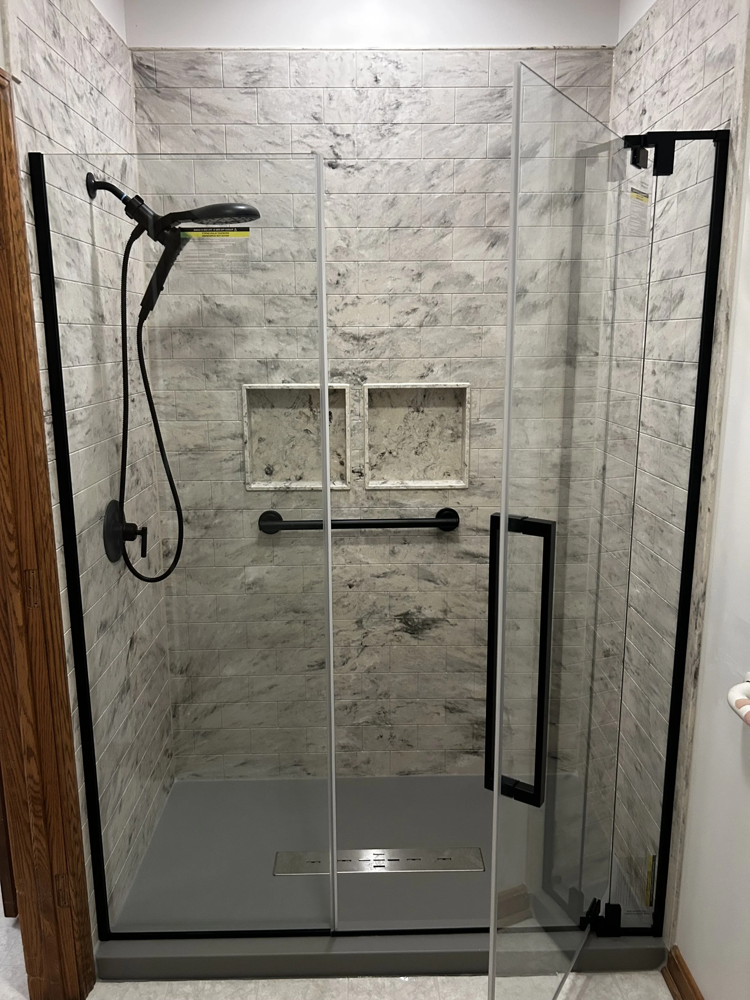 A walk-in shower with glass doors, beige marble tiles, a handheld showerhead, and two recessed shelves in the wall for toiletries.