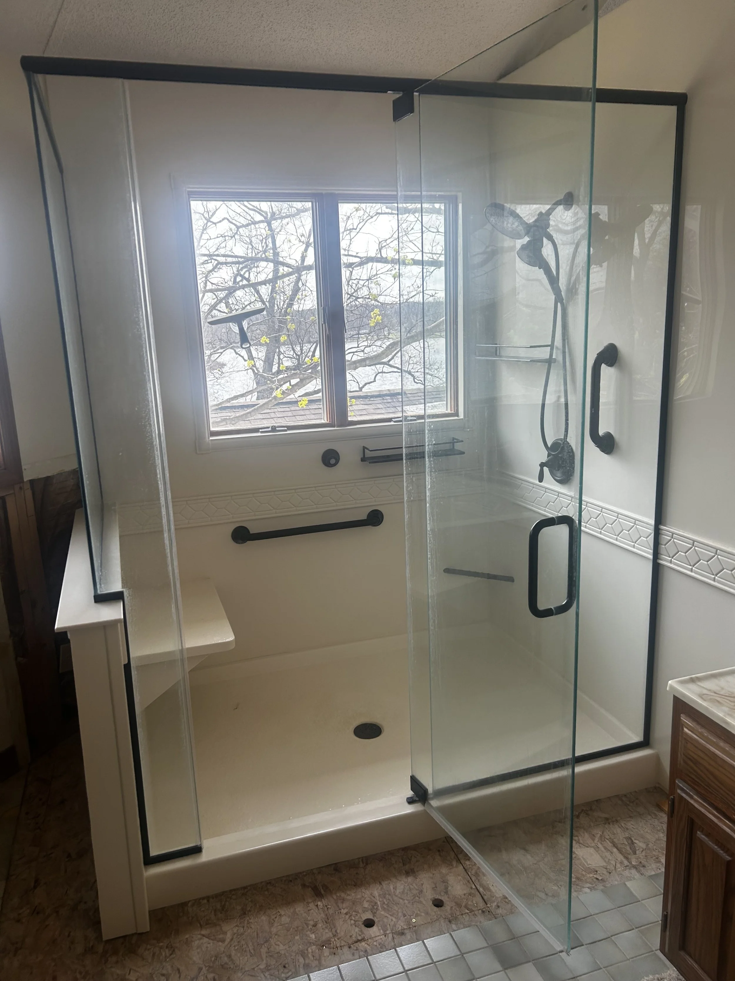 Bathroom shower with glass enclosure, built-in bench, black grab bars, and a window showing a tree outside.