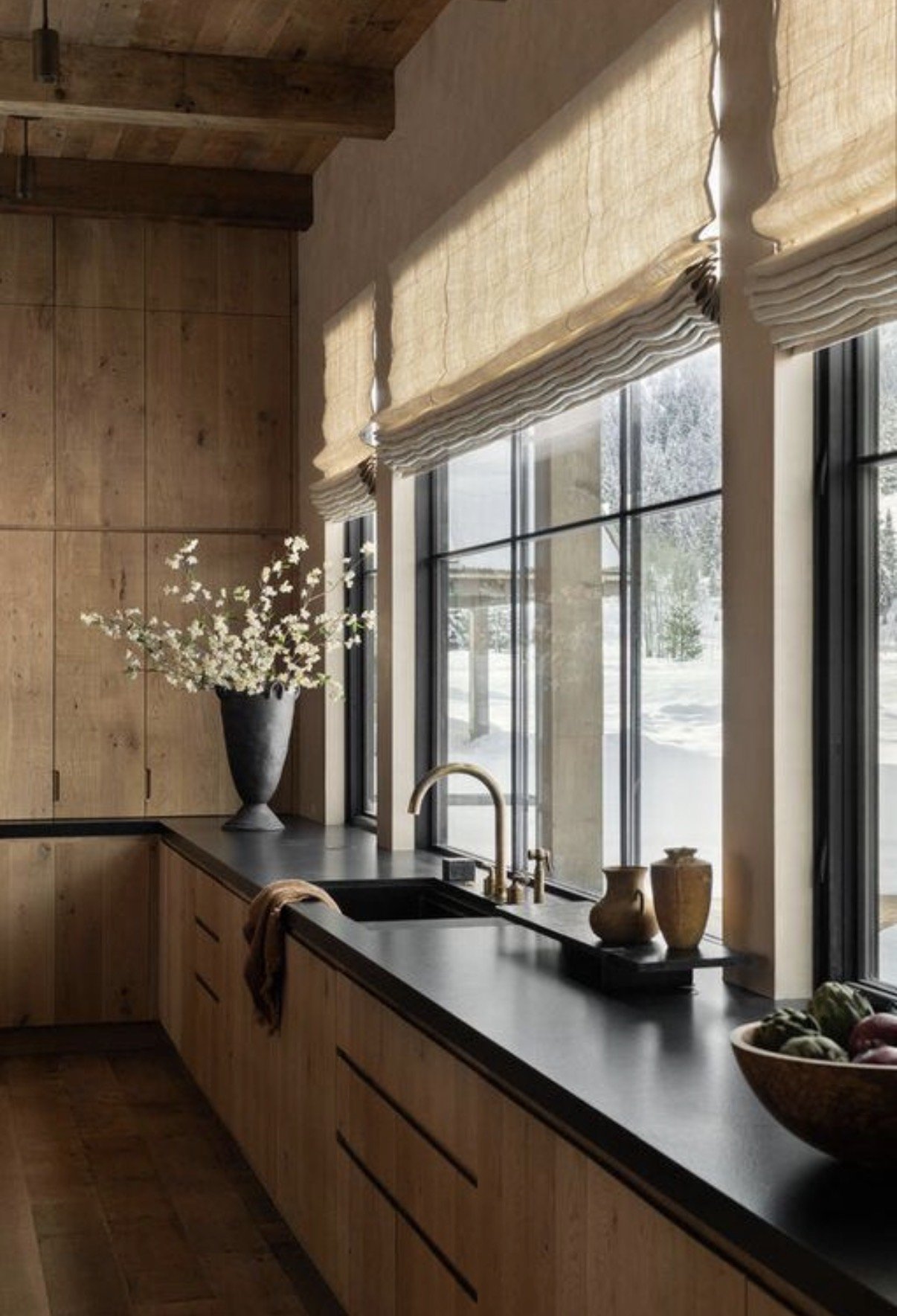 Kitchen with wooden cabinets, black countertop, and large window with beige roman shades. There is a black vase with white flowers, a bowl of fruit, and two small decorative vases on the counter.