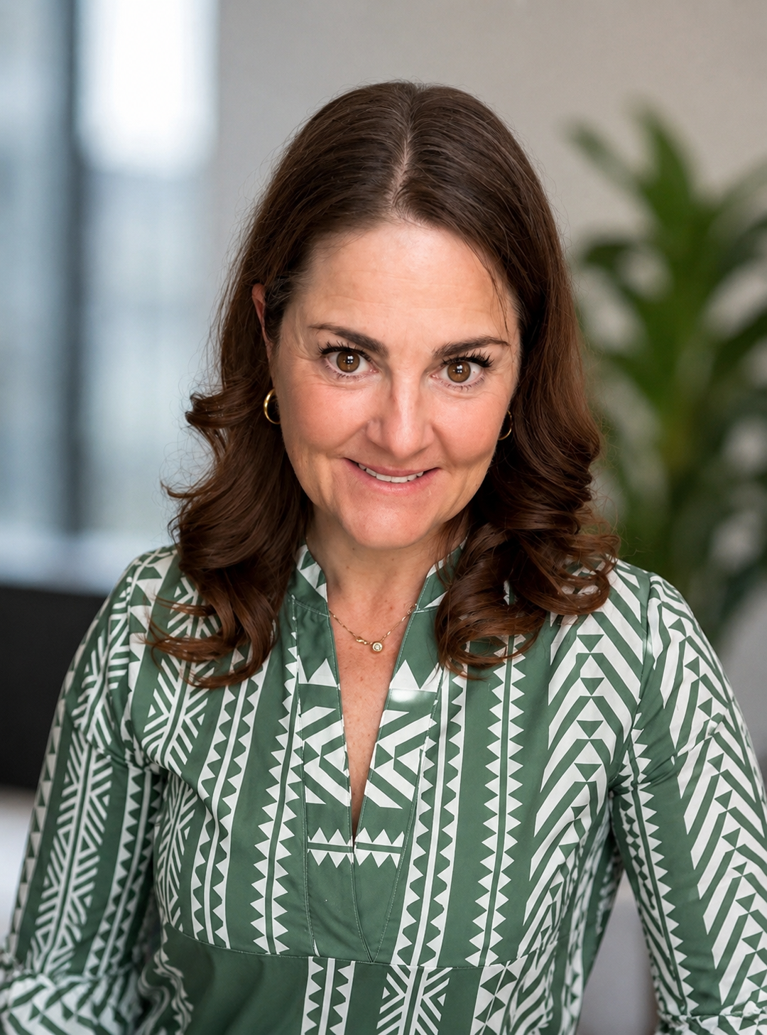 A woman with brown wavy hair and gold hoop earrings, wearing a green and white patterned blouse, smiling and looking at the camera in an office setting with a blurred green plant in the background.
