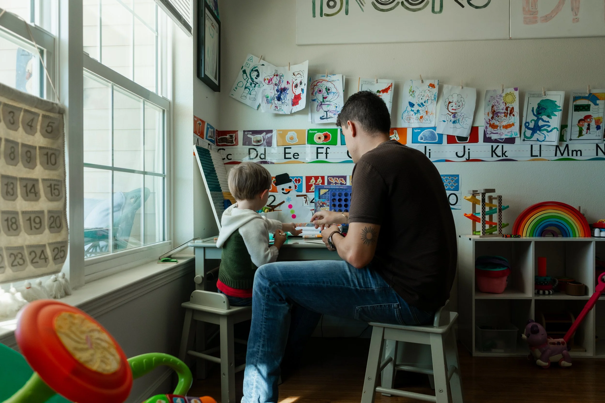 A man and a young boy sit at a small desk in a brightly lit room, working on arts and crafts. The room is decorated with colorful drawings hung on a string and educational posters, with a window to the left allowing natural light to fill the space.