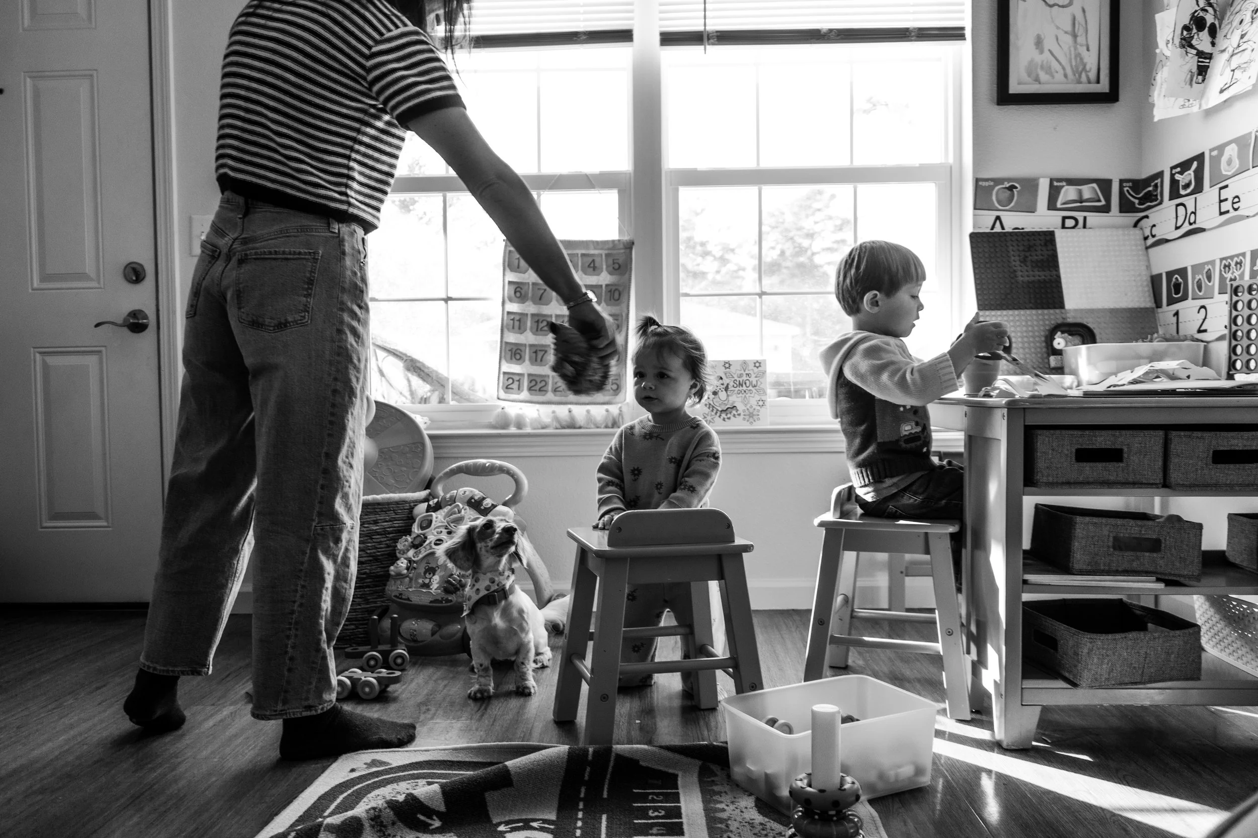 A woman in striped shirt and jeans standing in a child's playroom with two children, one girl and one boy, and a small dog. The girl sits on a small chair, and the boy is at a table. Toys, artwork, and educational materials are visible in the backgro