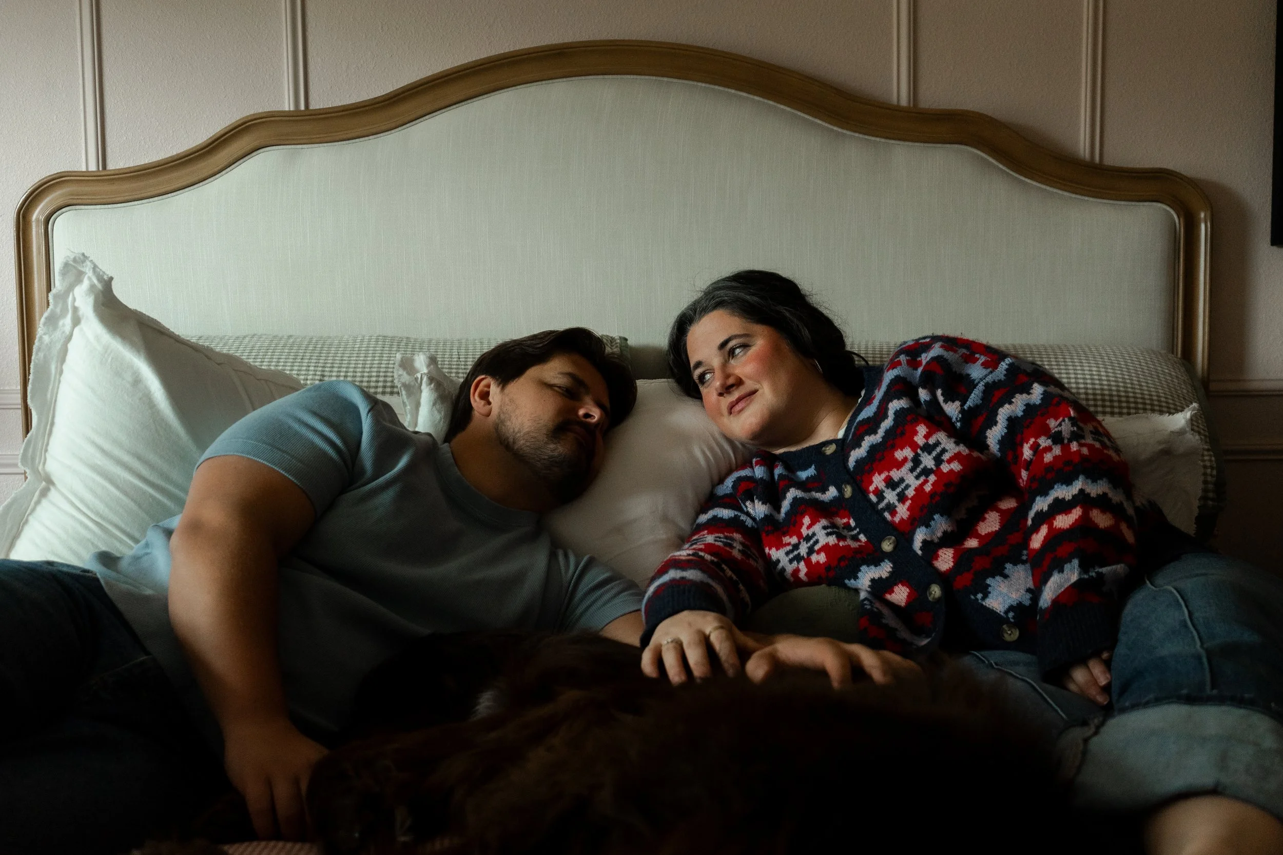 A man and woman lying on a bed, facing each other, with a dog partially visible in the foreground.