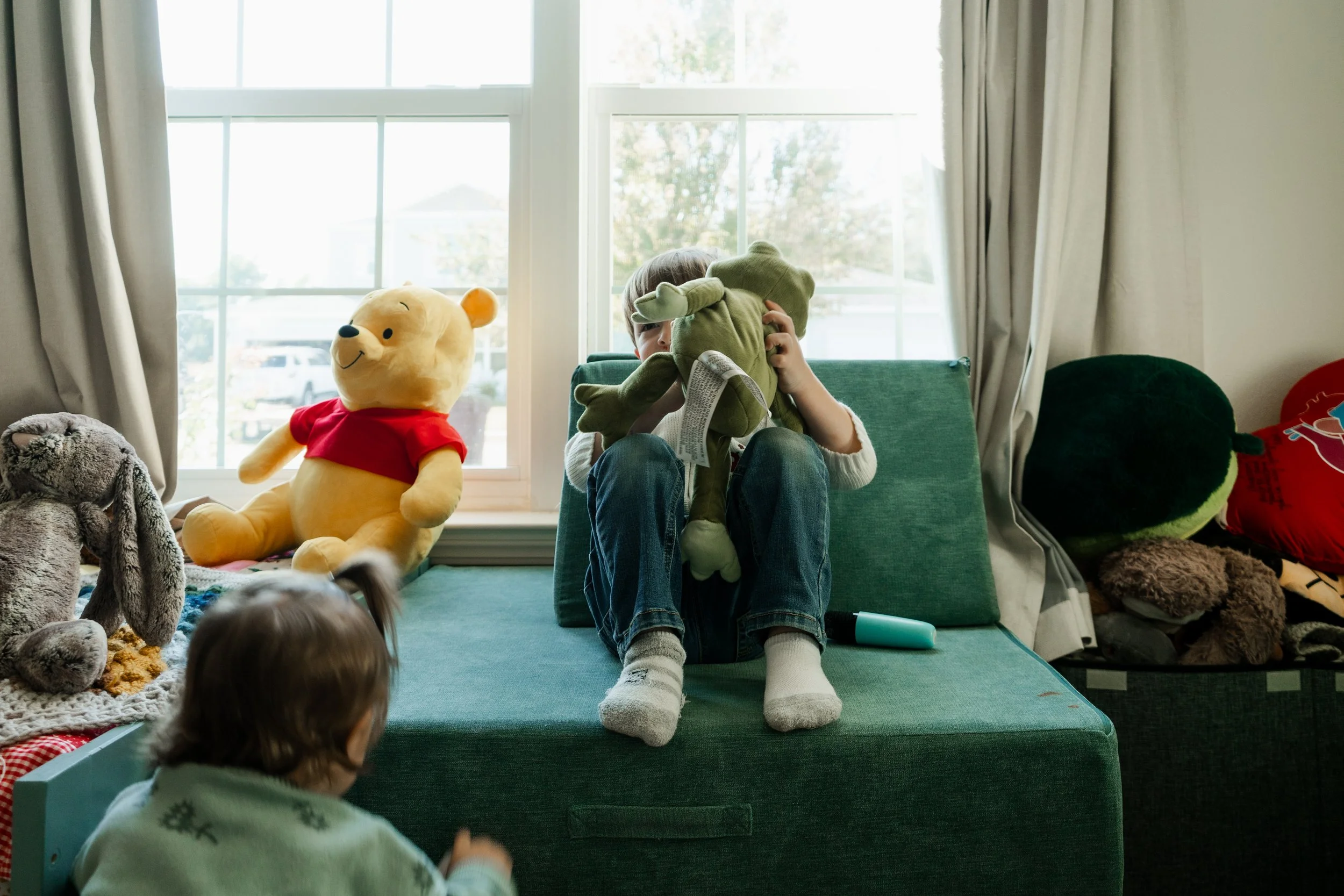 A young boy sitting on a green bed holding a plush frog toy in front of his face, with a window behind him. A girl is in the foreground looking at him. There are stuffed animals on the bed and around it.