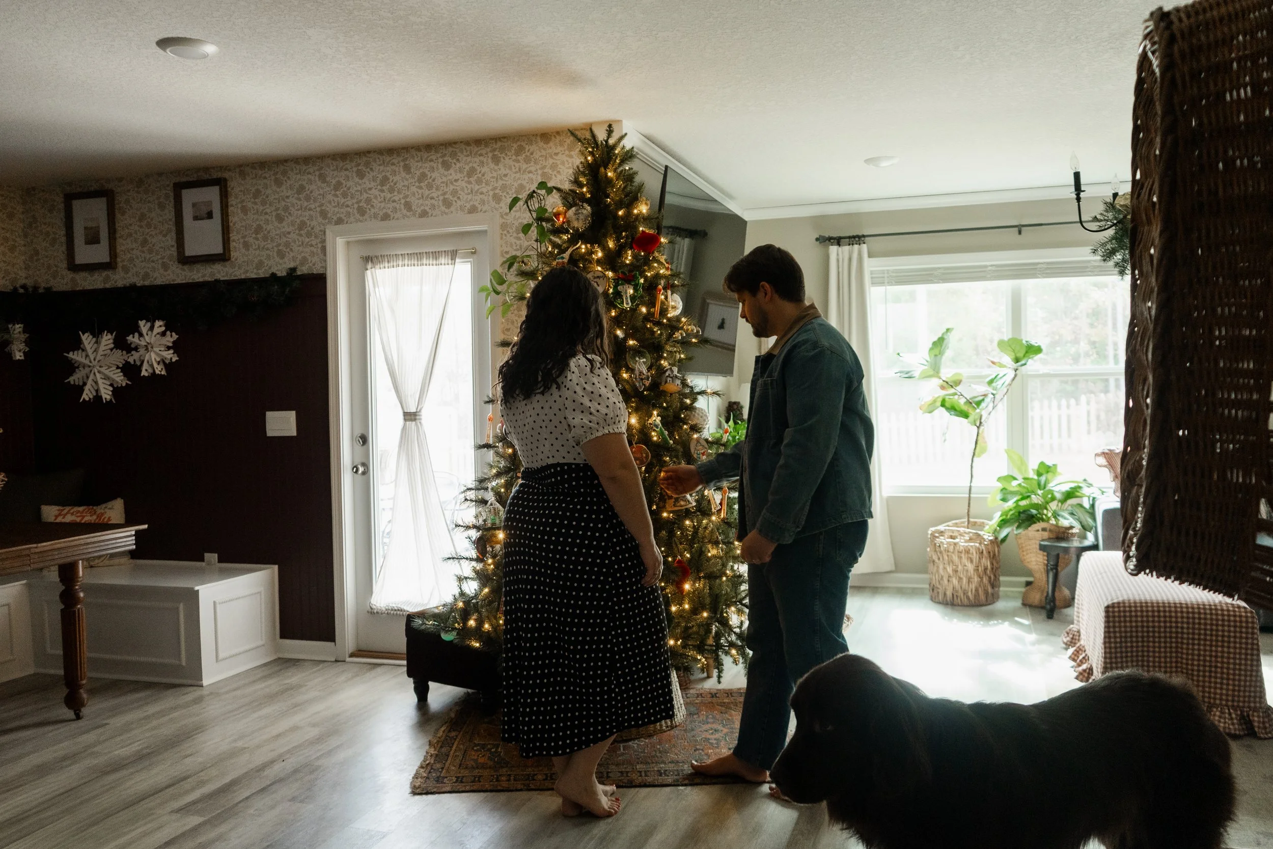 A man is giving a woman a ring in front of a decorated Christmas tree inside a home, with a black dog nearby.