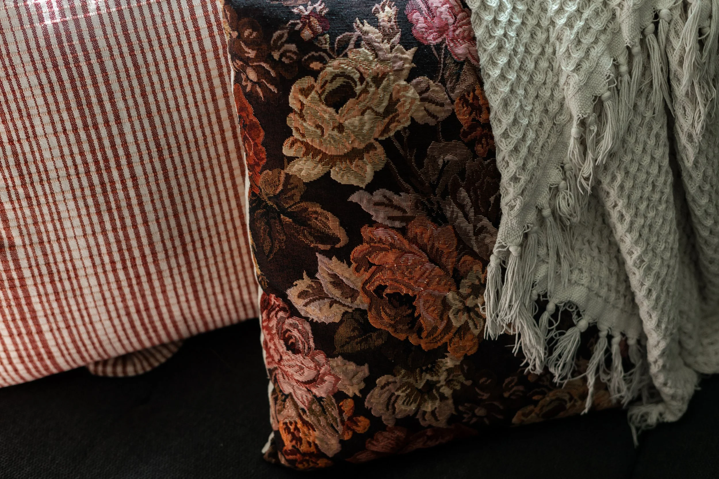 Close-up of two decorative pillows on a dark surface, one with a red and cream checkered pattern and the other with a floral tapestry design in dark colors with pink, peach, and cream flowers, partially covered by a light-colored textured blanket wit