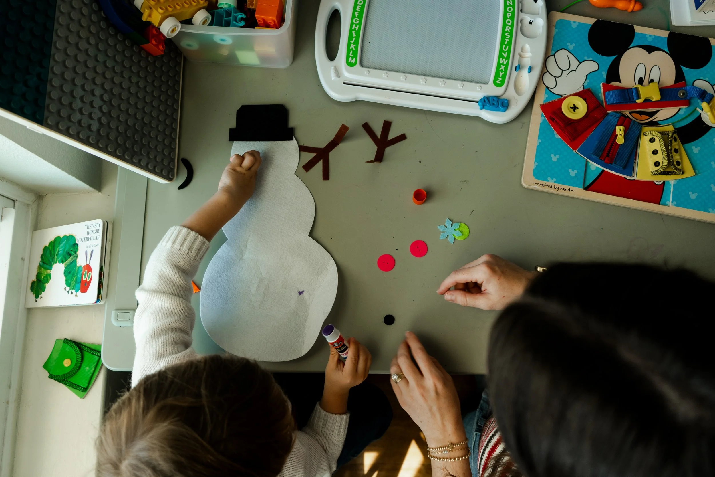 Two children and an adult are sitting at a table decorating a paper snowman craft with felt, buttons, and other craft supplies. The table has various craft items, books, and a whiteboard nearby.