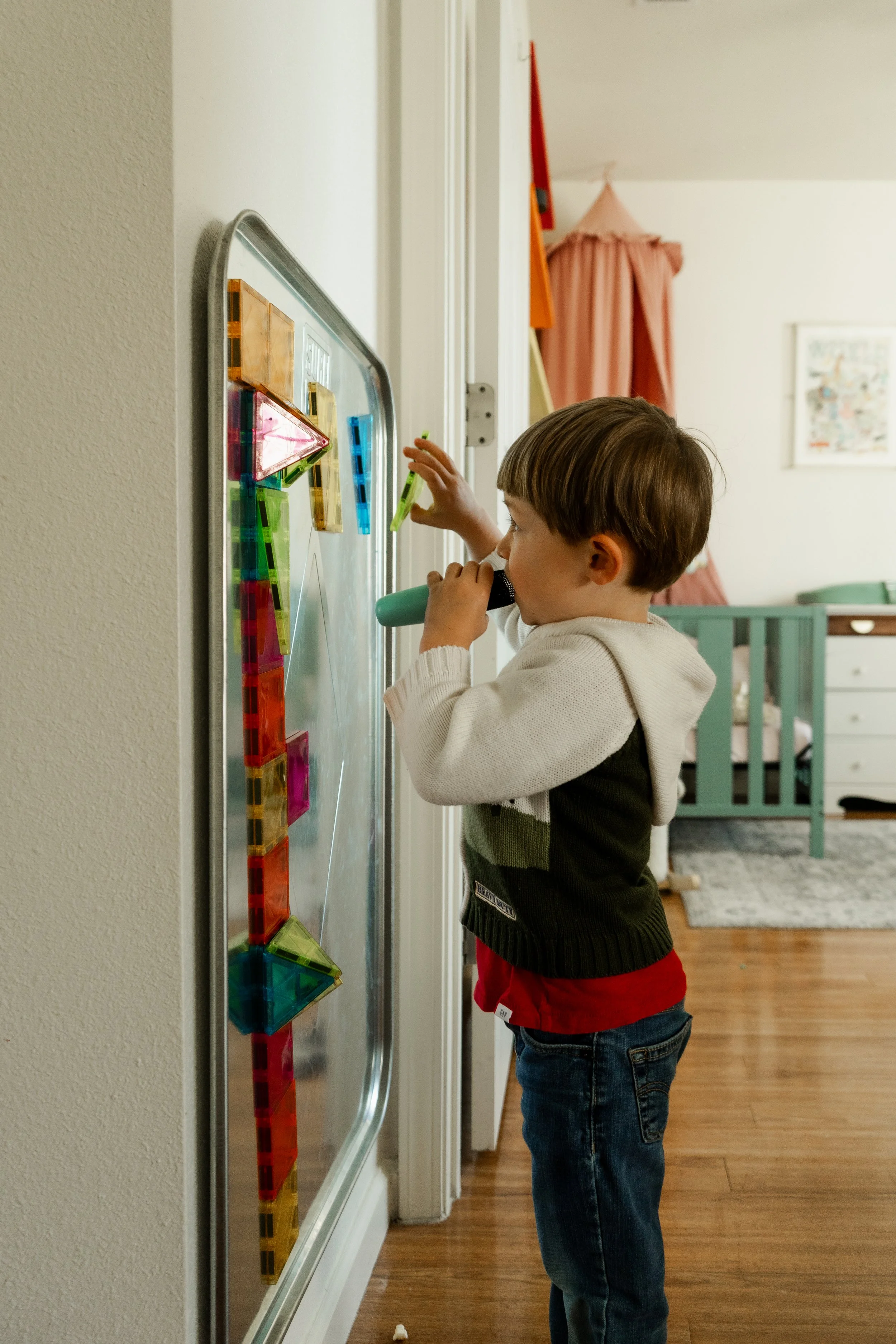 Young boy looking through a magnifying glass at magnetic building tiles on a magnetic board in a child's room.