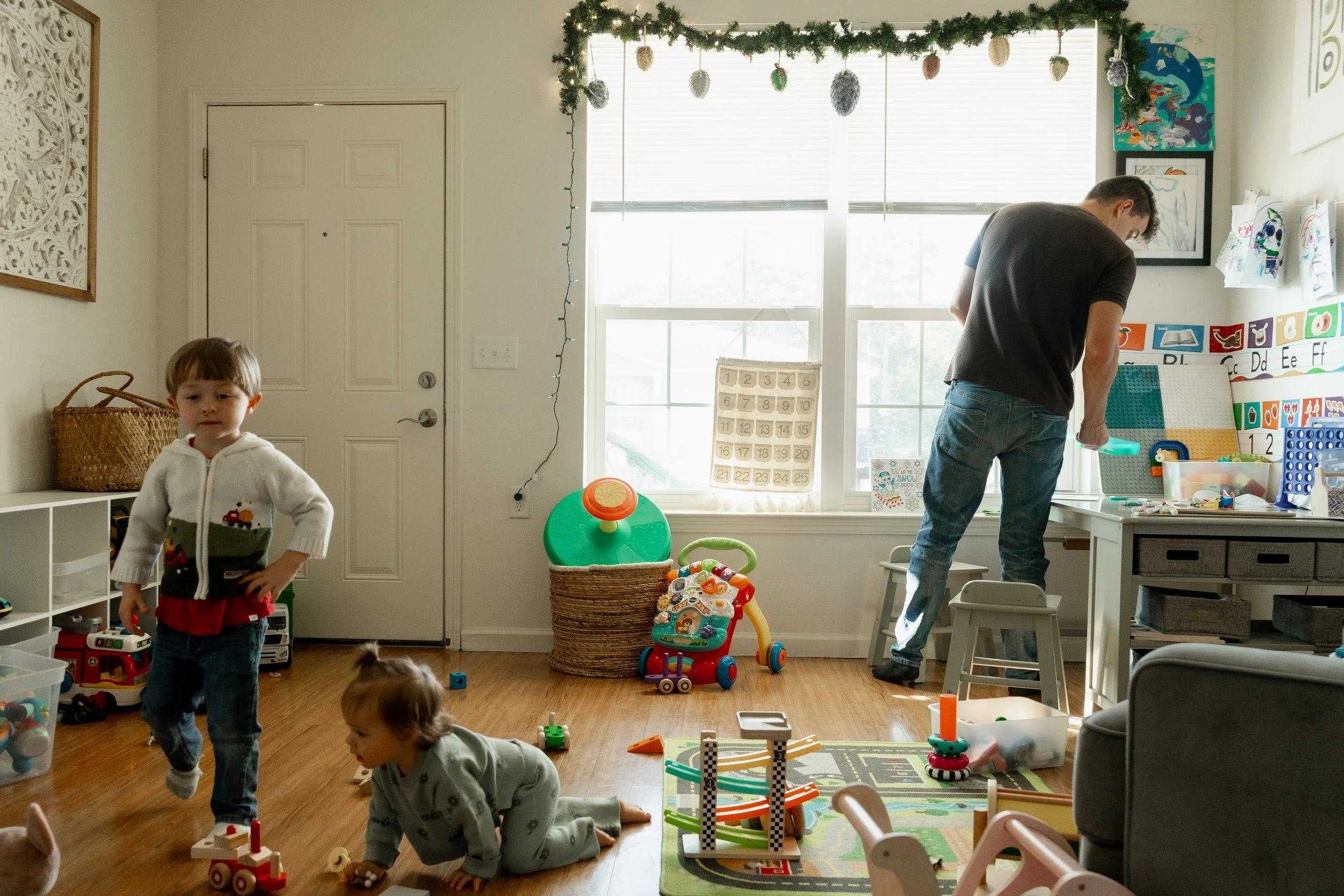 A room filled with toys, children playing on the floor, a man working at a desk near a large window decorated with hanging garland and handmade ornaments, children engaging with toys and puzzles on the floor.