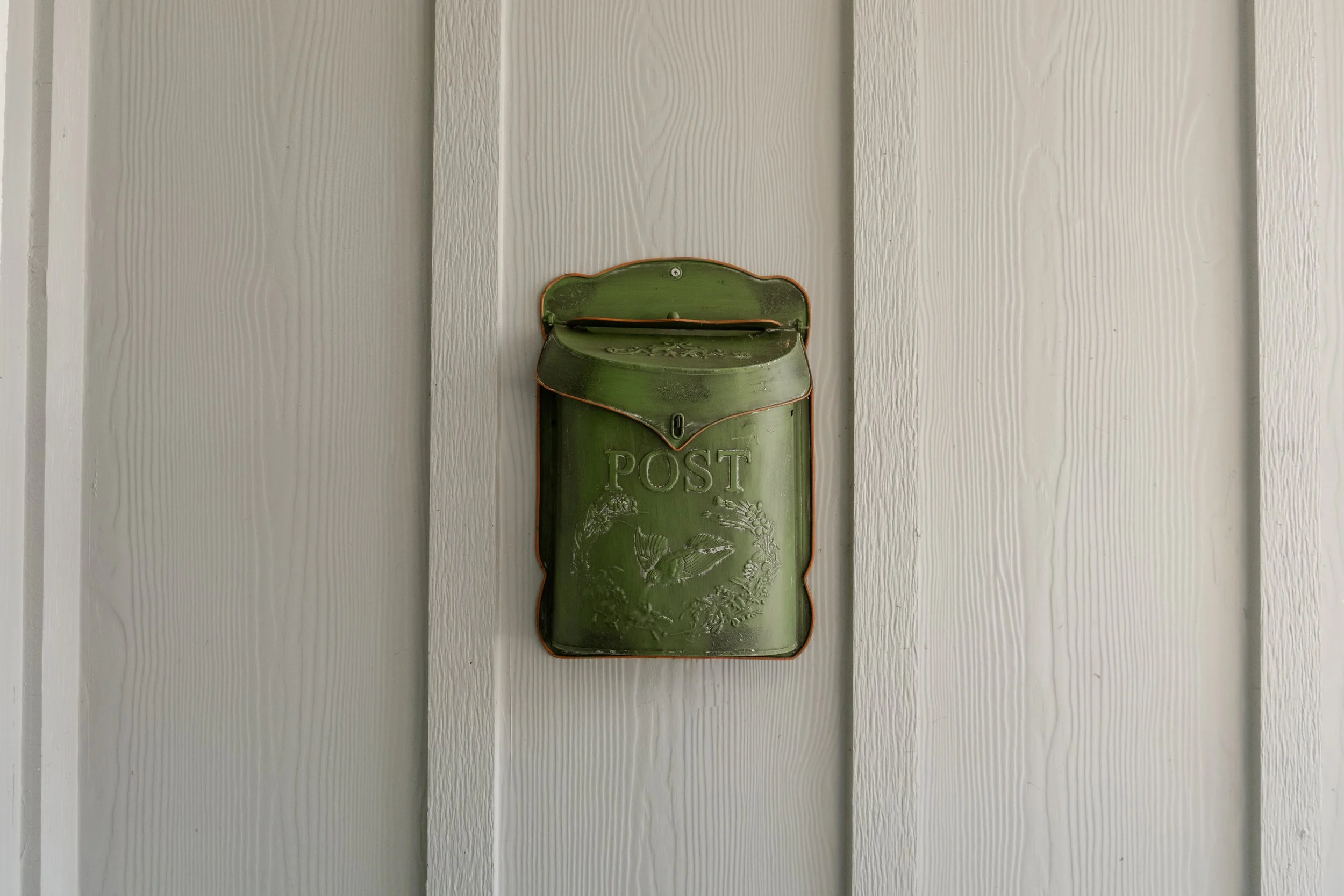 Green vintage metal mailbox with the word POST on it, decorated with a bird and foliage design, mounted on a white wooden wall.