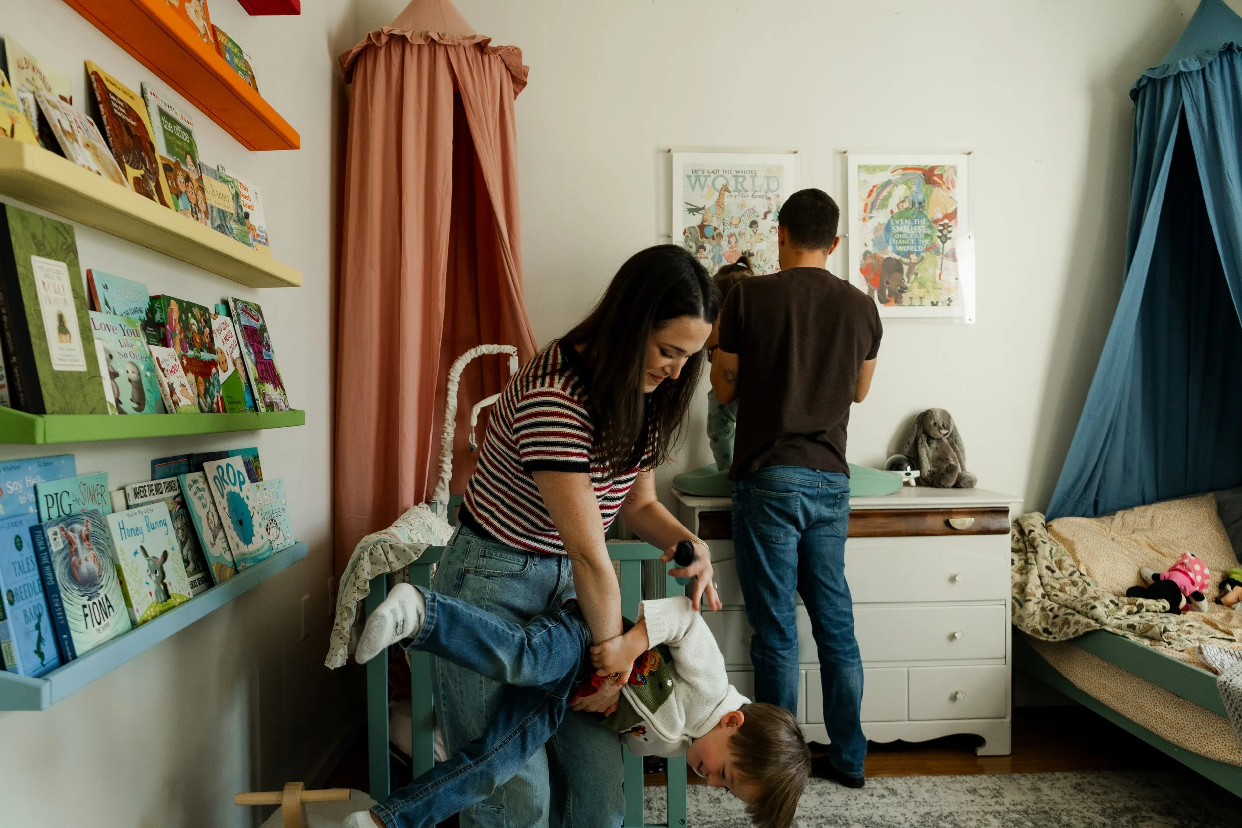 A woman is playfully hanging upside down over a child's lap in a cozy, book-filled child's room, with two other people in the background, one standing at a changing table and another near a bed with stuffed animals.