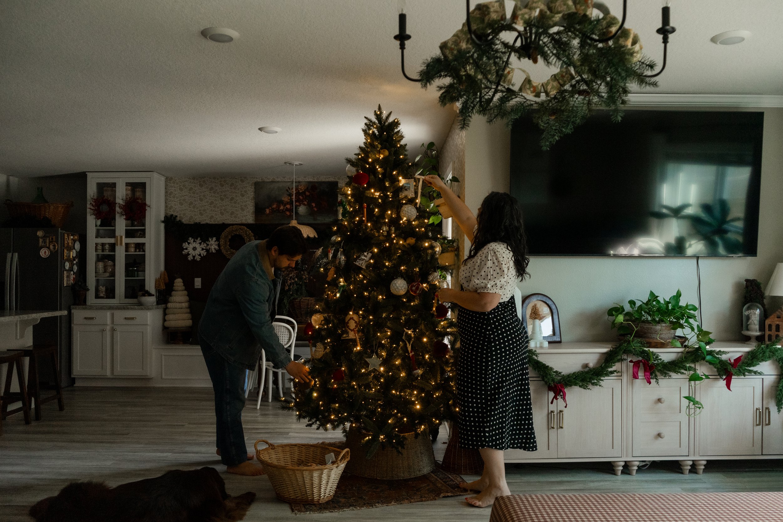 A man and a woman decorating a Christmas tree in a living room, with Christmas lights and ornaments. The room is decorated for the holidays with garlands, wreaths, and presents. A large flat-screen TV is mounted on the wall, and there are plants and 