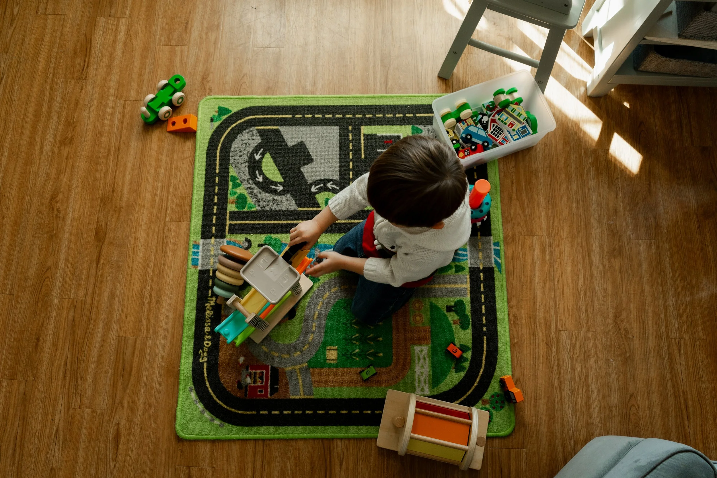 A young child sitting on a colorful playmat with a road and town design, playing with various toy transportation vehicles and blocks. Sunlight streams in from the right side, illuminating the scene.