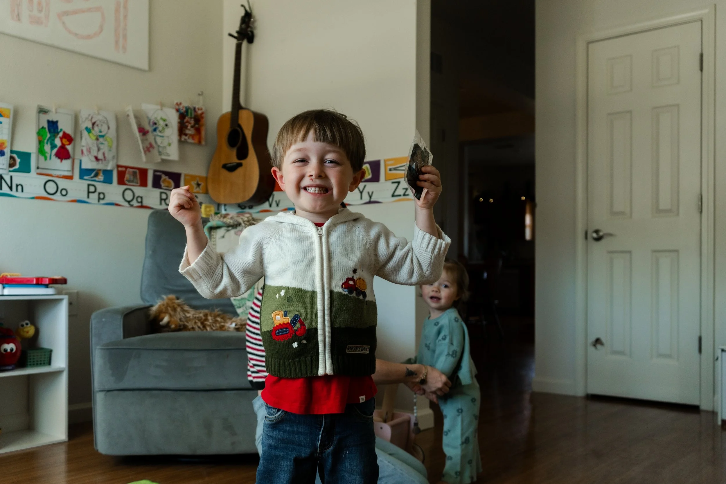 Smiling young boy in a cozy living room holding a small bag in one hand and raising his other fist, with a younger child in pajamas and a parent's hand in the background. The room has a guitar, children's artwork, and educational alphabet decorations