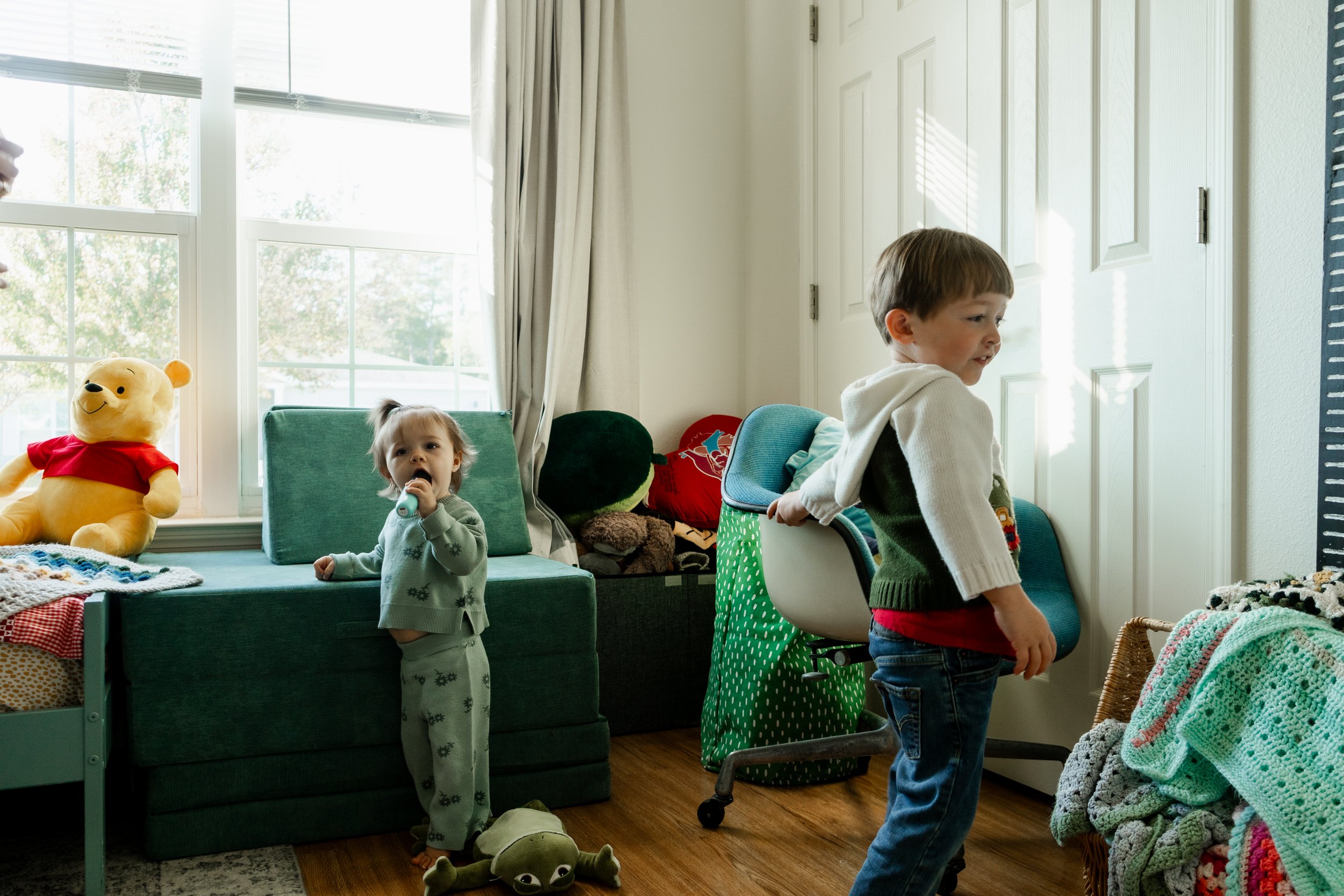 Two children in a brightly lit room with large windows, surrounded by toys and blankets. A young girl stands near a green cushioned bench holding a toy, while an older boy stands beside a desk cluttered with blankets and stuffed animals.