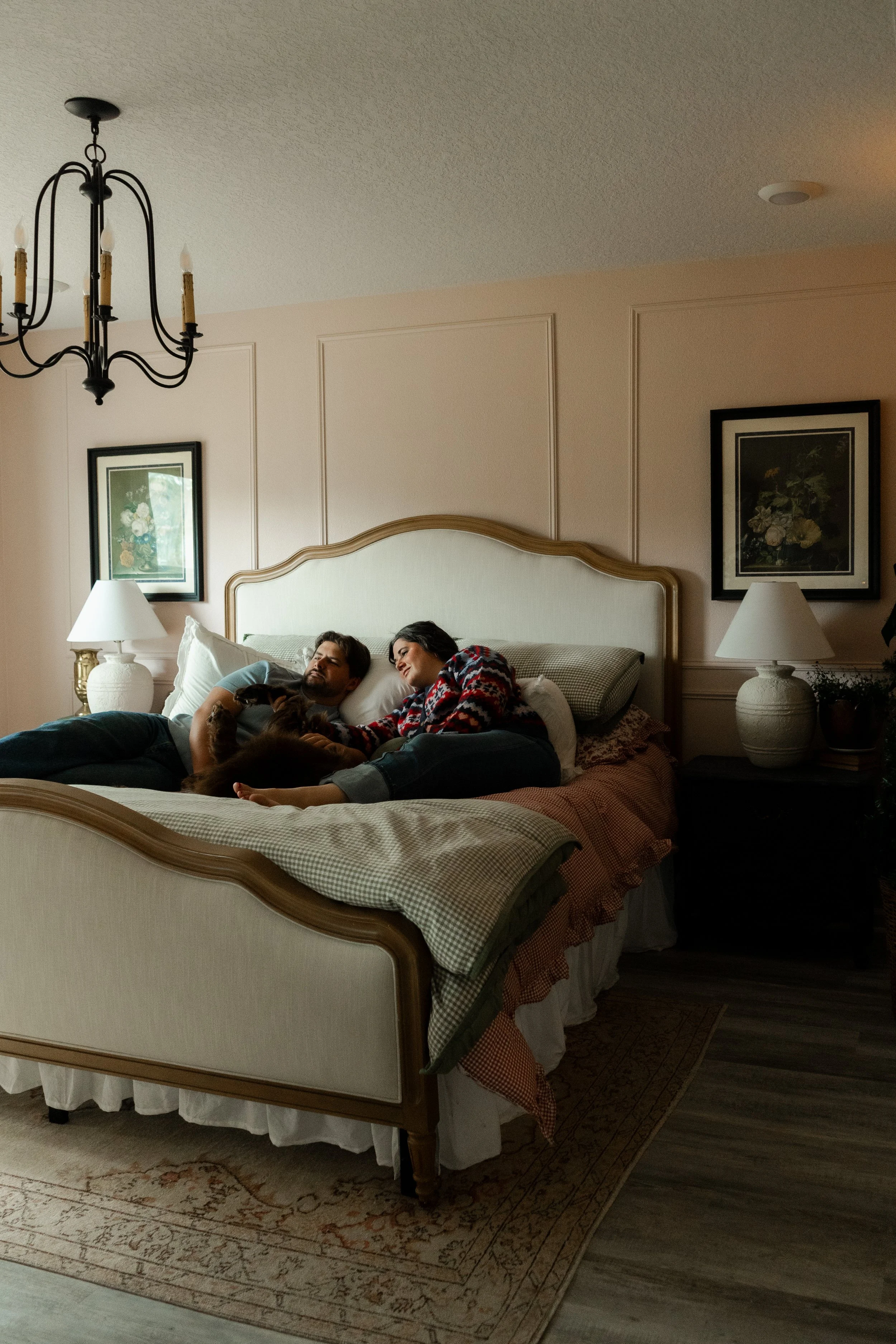 Two people lying on a bed, cuddling with a dog in a cozy bedroom with bedside lamps, framed pictures, and a chandelier.