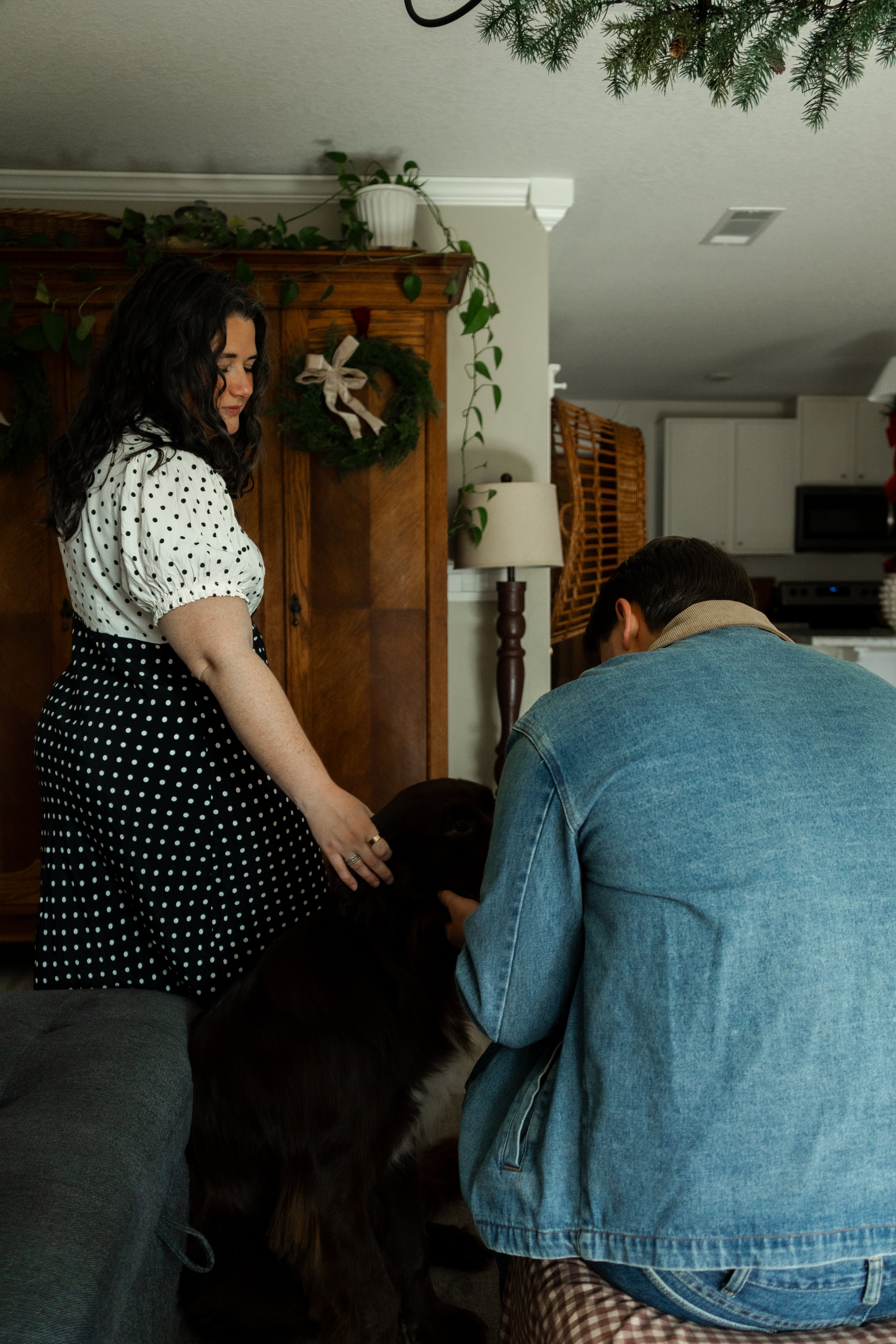 A woman and a man are interacting with a black dog in a cozy living room decorated for the holidays.