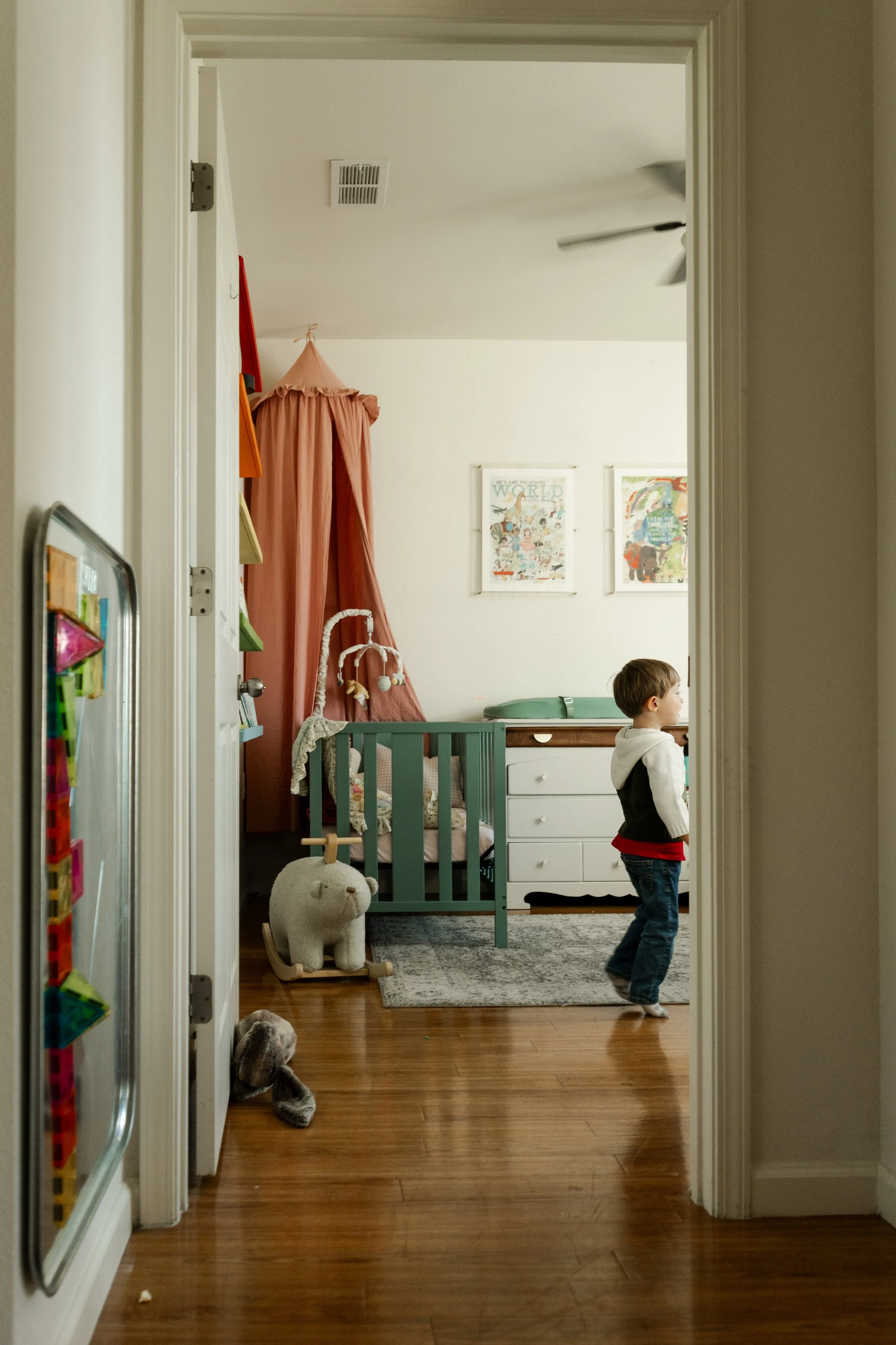 A young boy in a bedroom, looking to the right, with a green crib, colorful wall art, a pink canopy, plush toys, and a changing table.