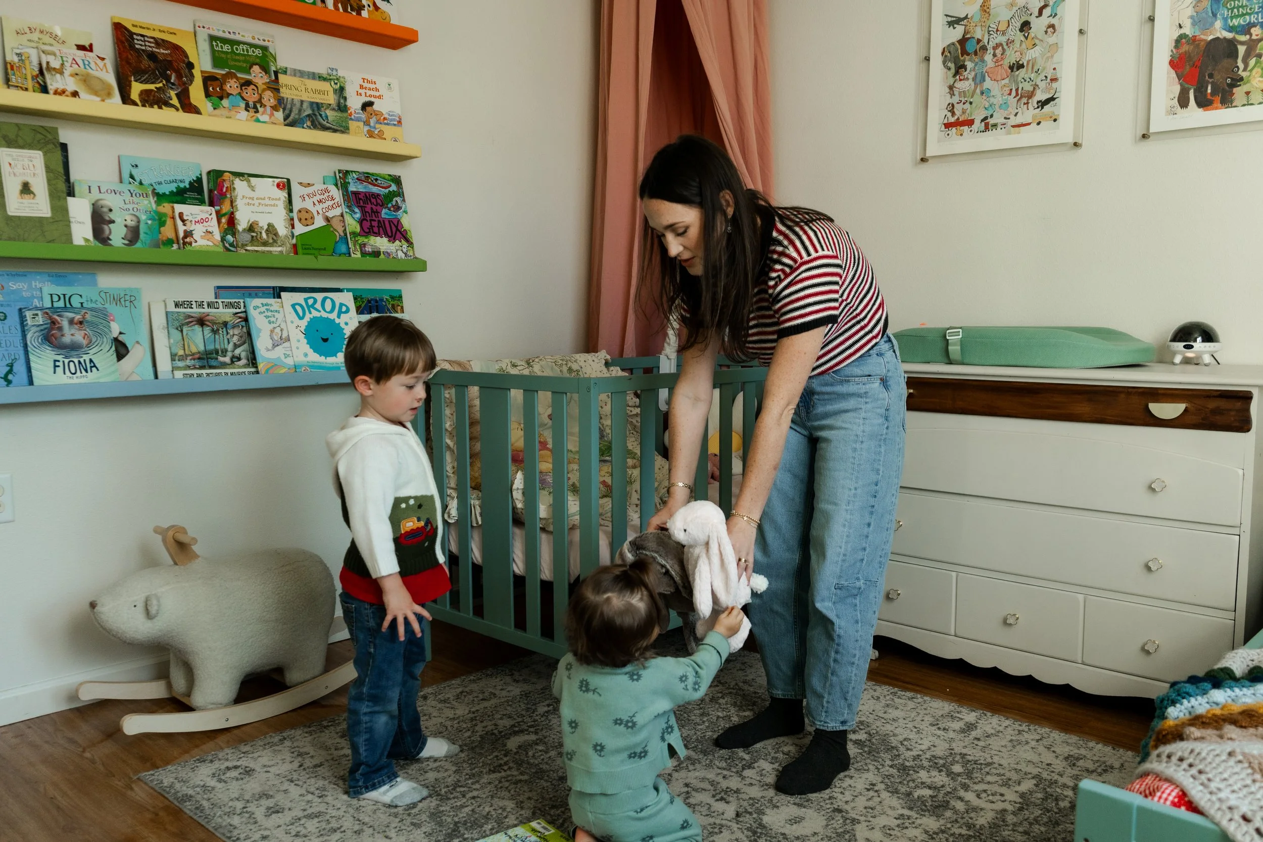A woman and two children in a nursery, with the woman holding a plush bunny, as the children hold the bunny's ears. The room has bookshelves with children's books, a wood rocking sheep, and colorful wall art.