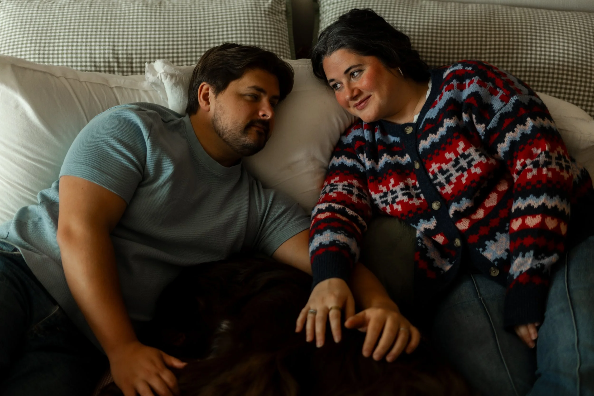 A man and woman relaxing on a bed, holding hands, with their heads close together, looking into each other's eyes.