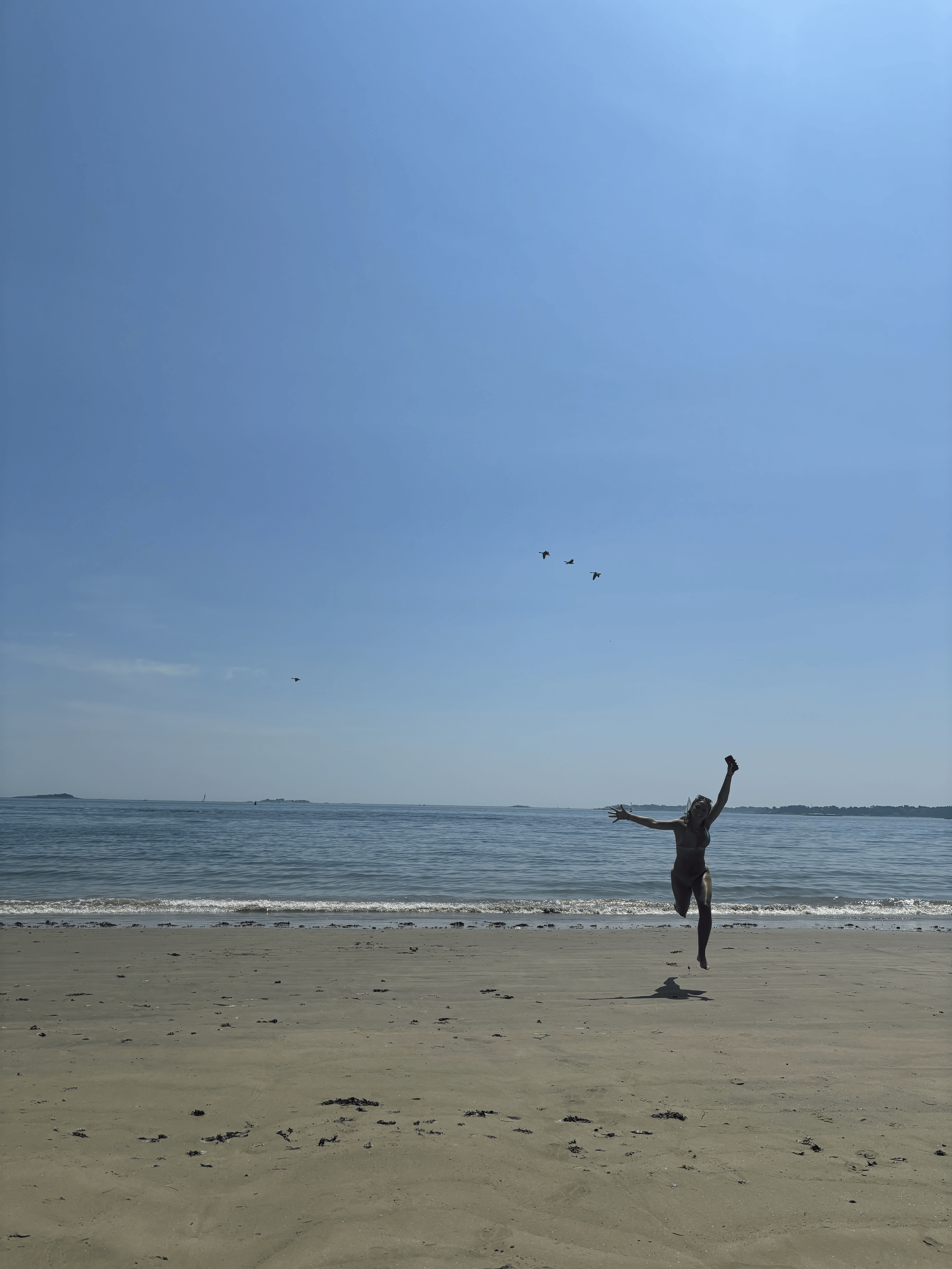 A woman in a swimsuit jumping on a sandy beach with the ocean in the background, small waves approaching the shore, a clear blue sky, and birds flying overhead.