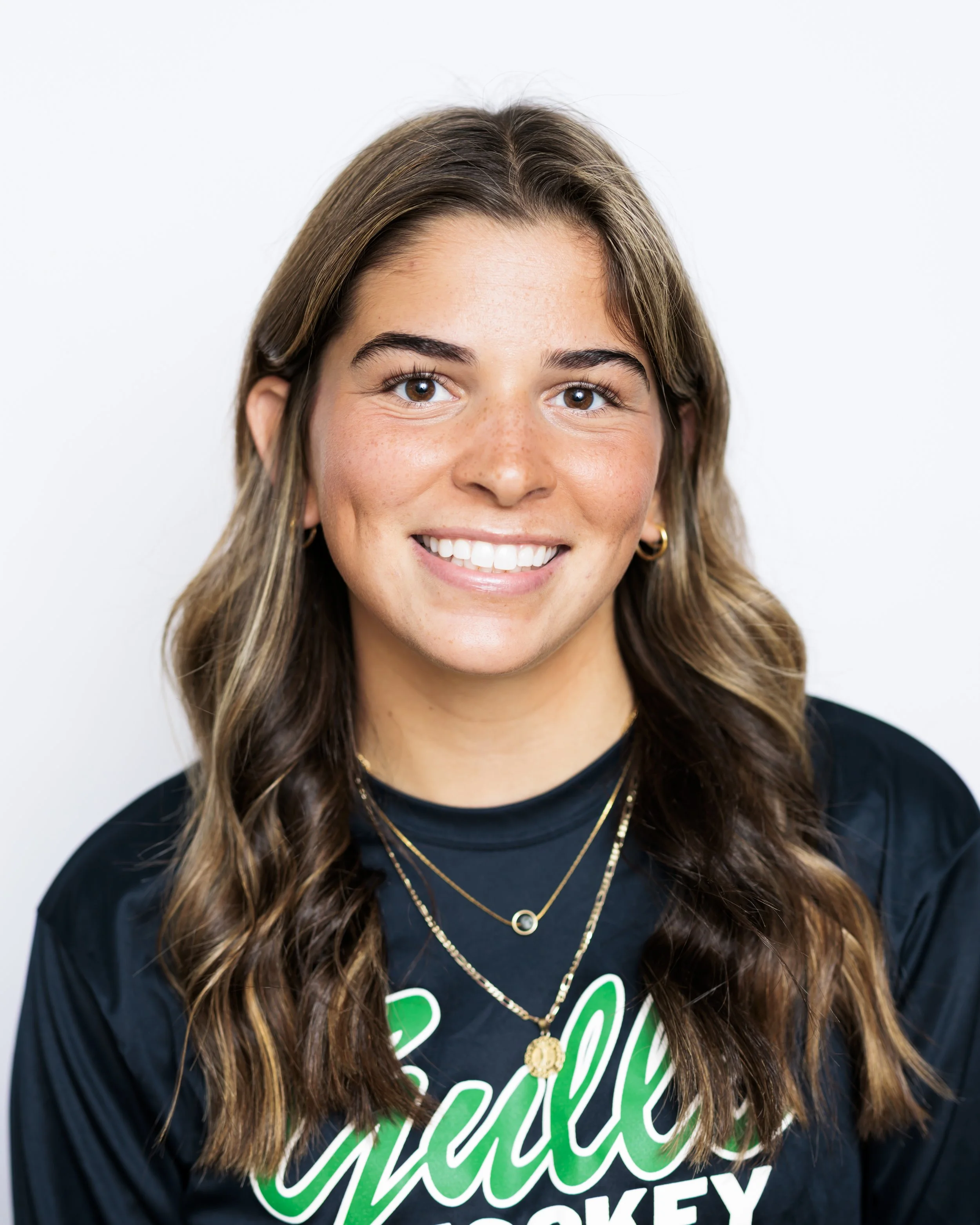 A young woman with brown hair, wearing layered necklaces, hoop earrings, and a black t-shirt with green and white text, smiling at the camera against a plain white background.