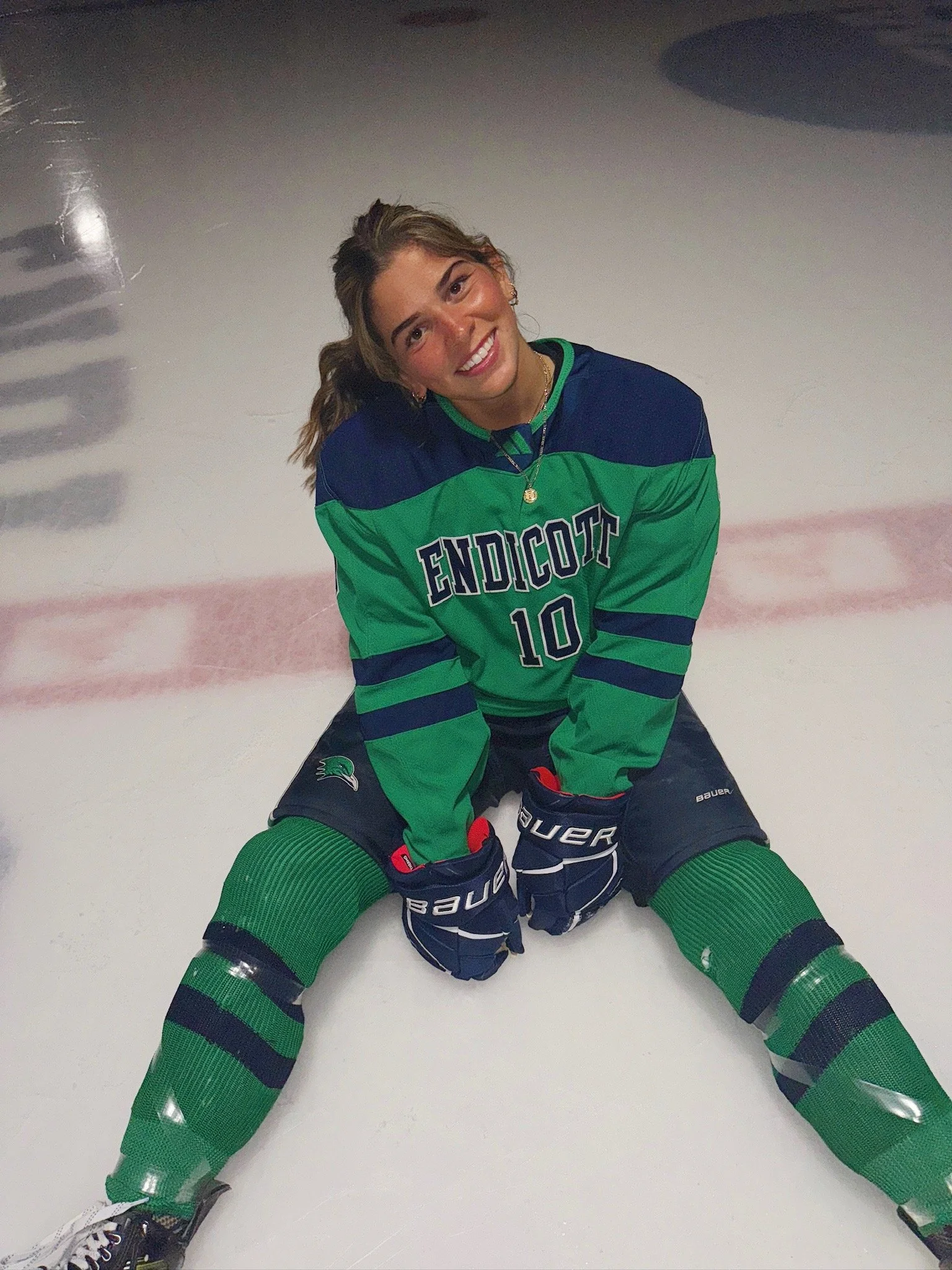 A young woman in a green and blue hockey uniform sitting on the ice rink with her legs spread out and smiling at the camera.