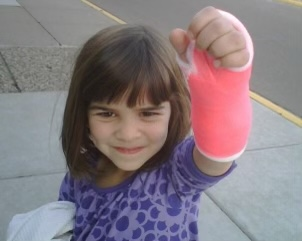 Young girl with a cast on her arm, smiling and holding it up outside on a sidewalk.