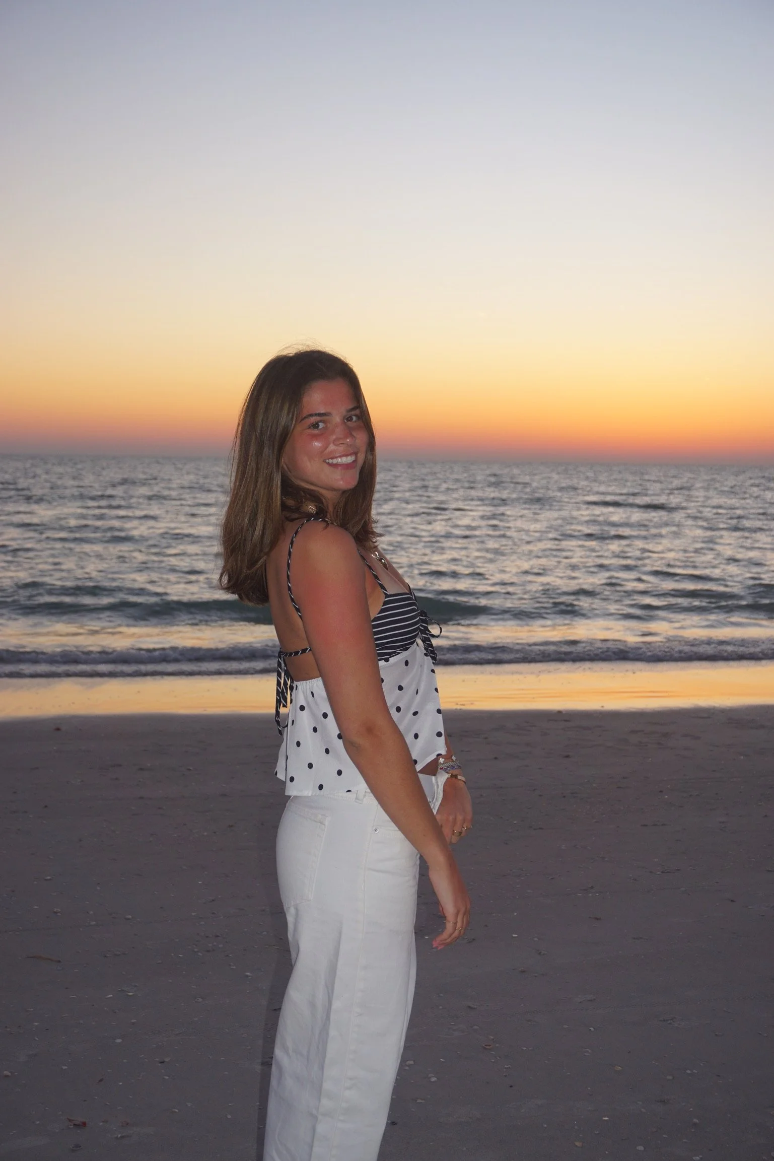 A woman with shoulder-length brown hair smiling on a beach during sunset, wearing a striped and polka dot sleeveless top and white pants.