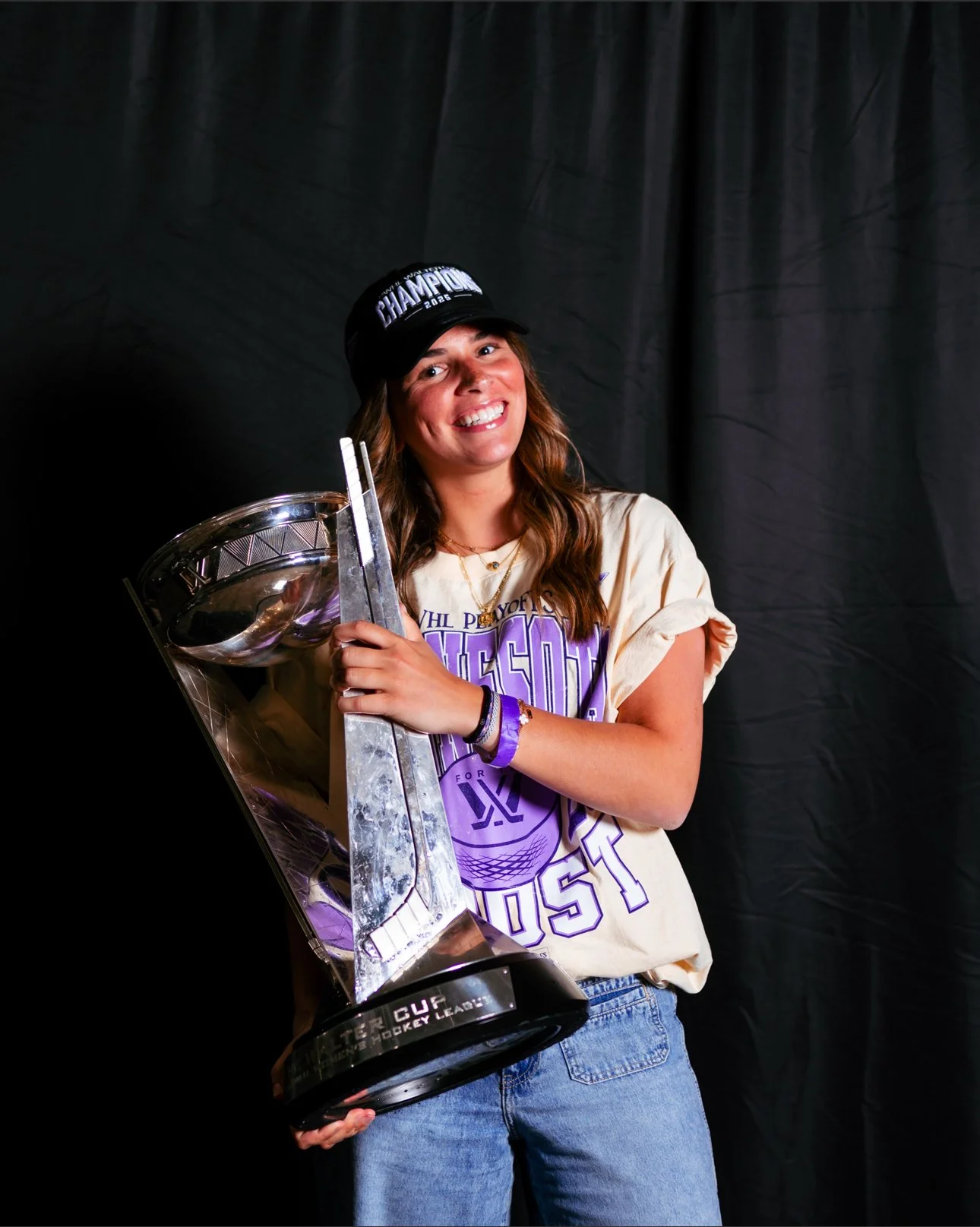 A woman dressed in a beige T-shirt and blue jeans, wearing a black cap that says 'Champion 2023', is holding a large silver hockey trophy with a big smile on her face, standing against a black backdrop.
