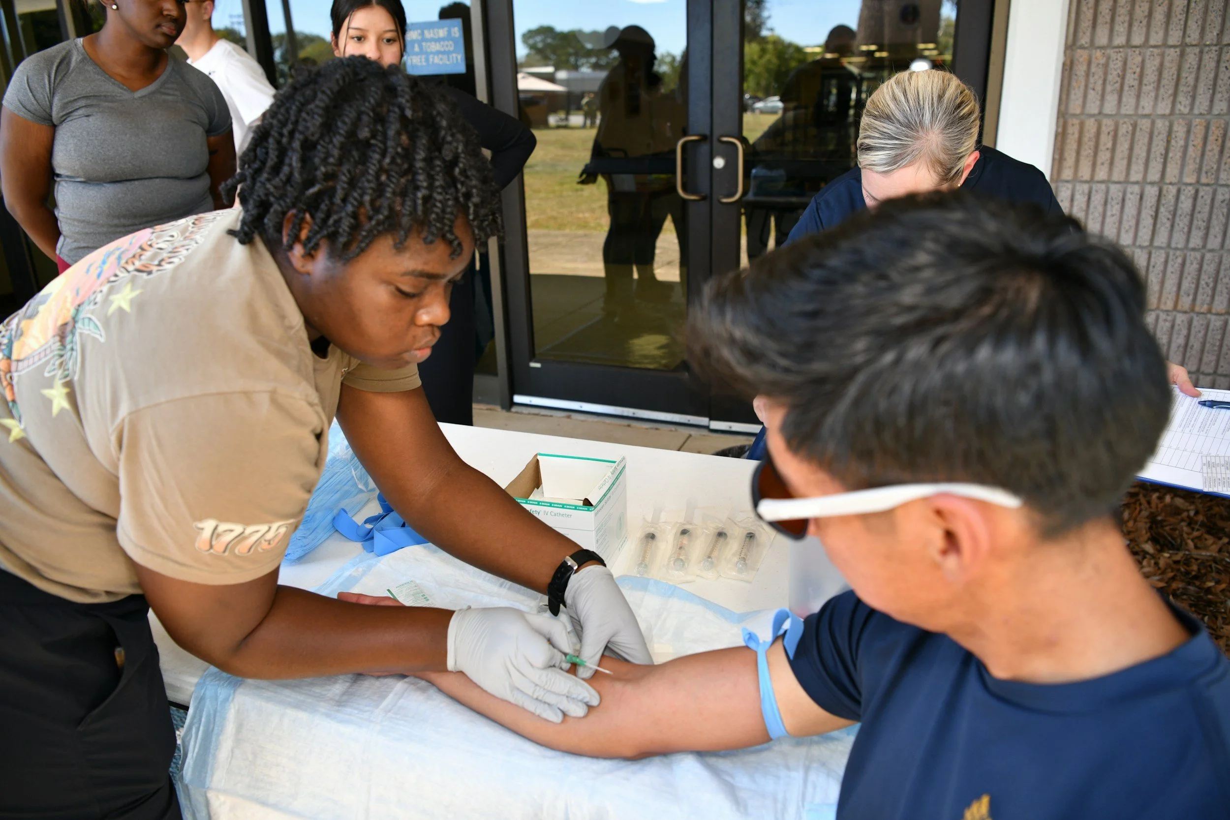 A person with curly hair and beige shirt drawing blood from a man's arm with a needle while other people observe.