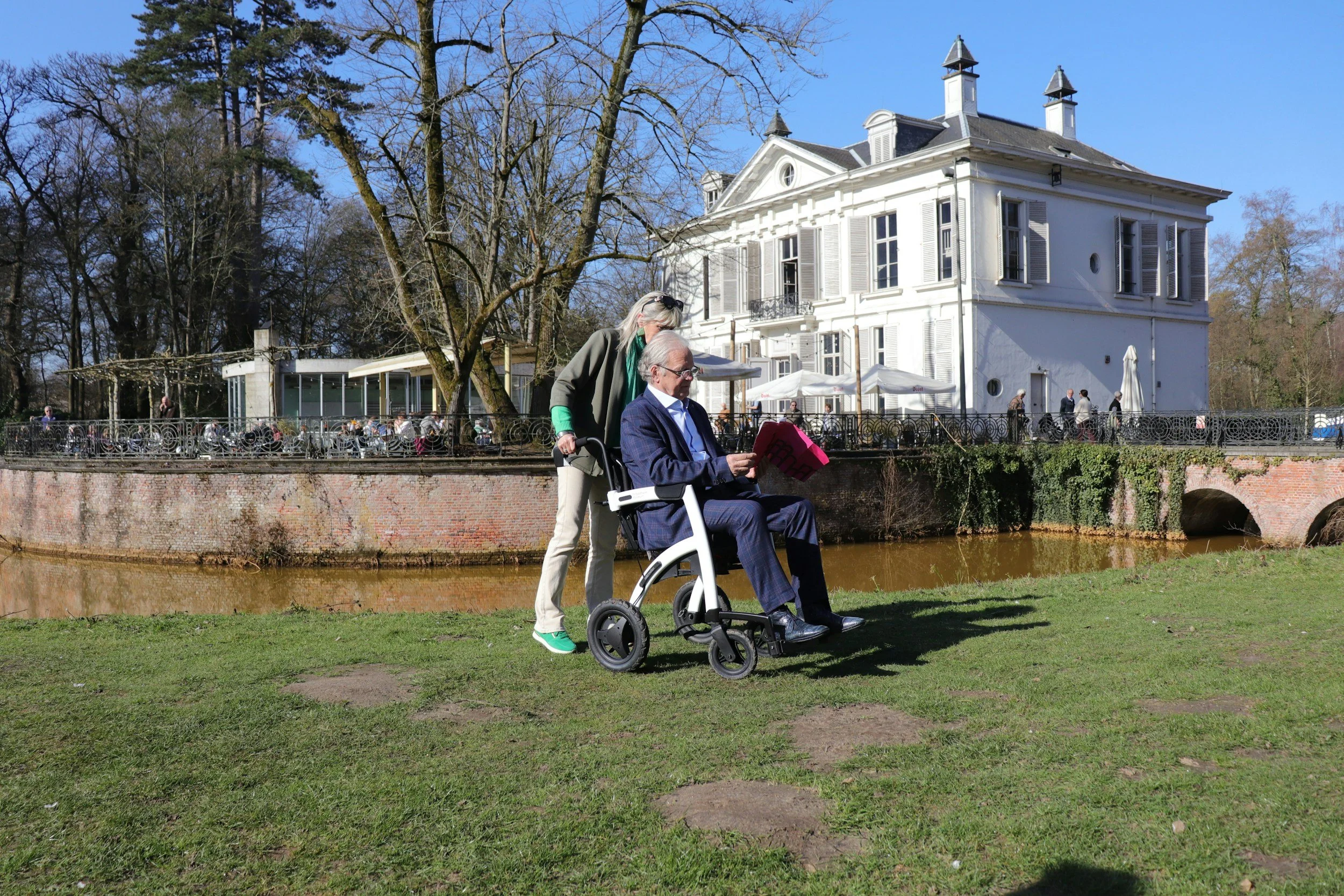 A man in a wheelchair and a woman standing behind him are outdoors on a grassy area near a river, with a large white house and trees in the background. The woman is reading a pink newspaper or magazine to the man.