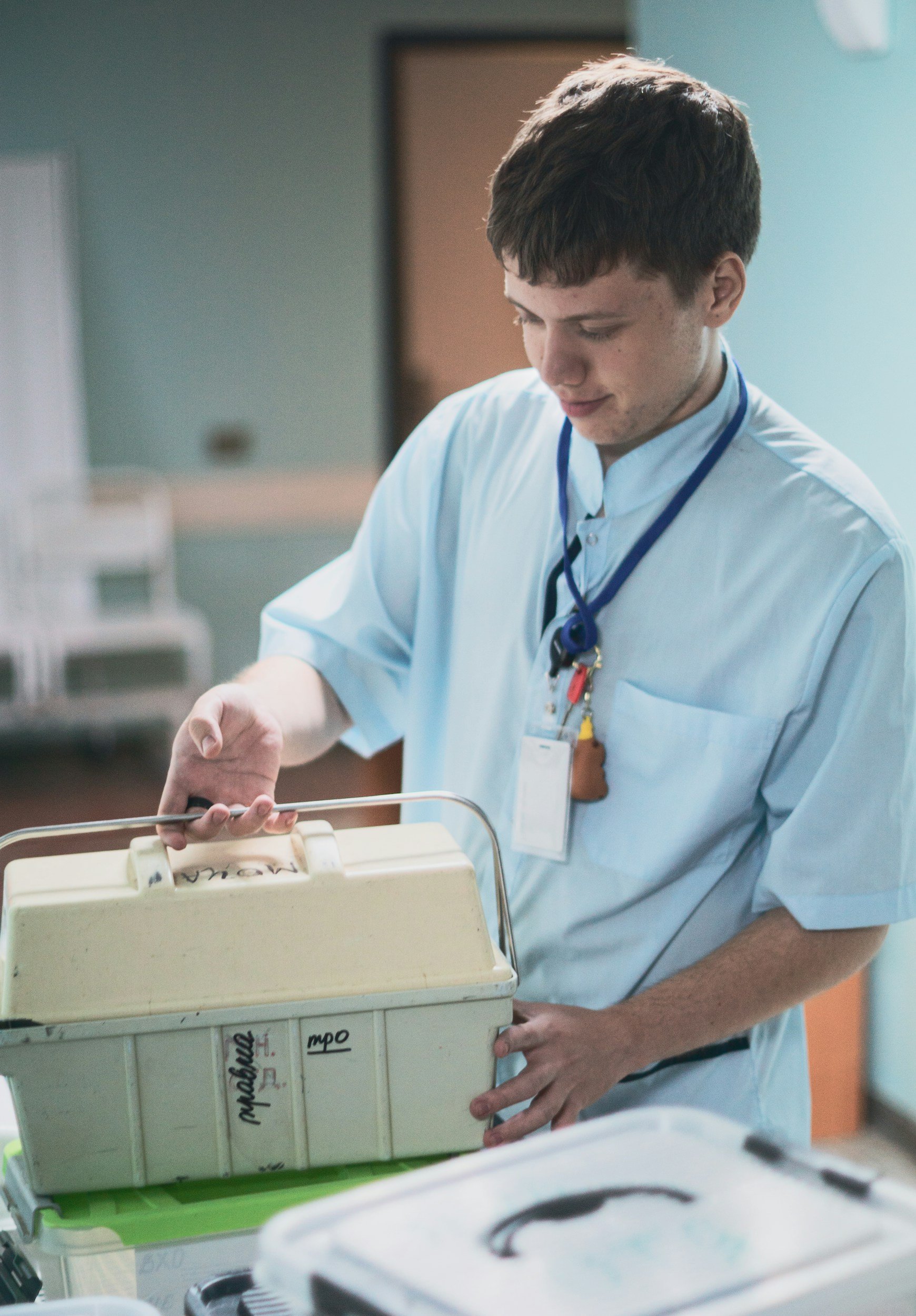 Young male nurse in light blue scrubs with a stethoscope around his neck, looking into a medical container in a hospital setting.