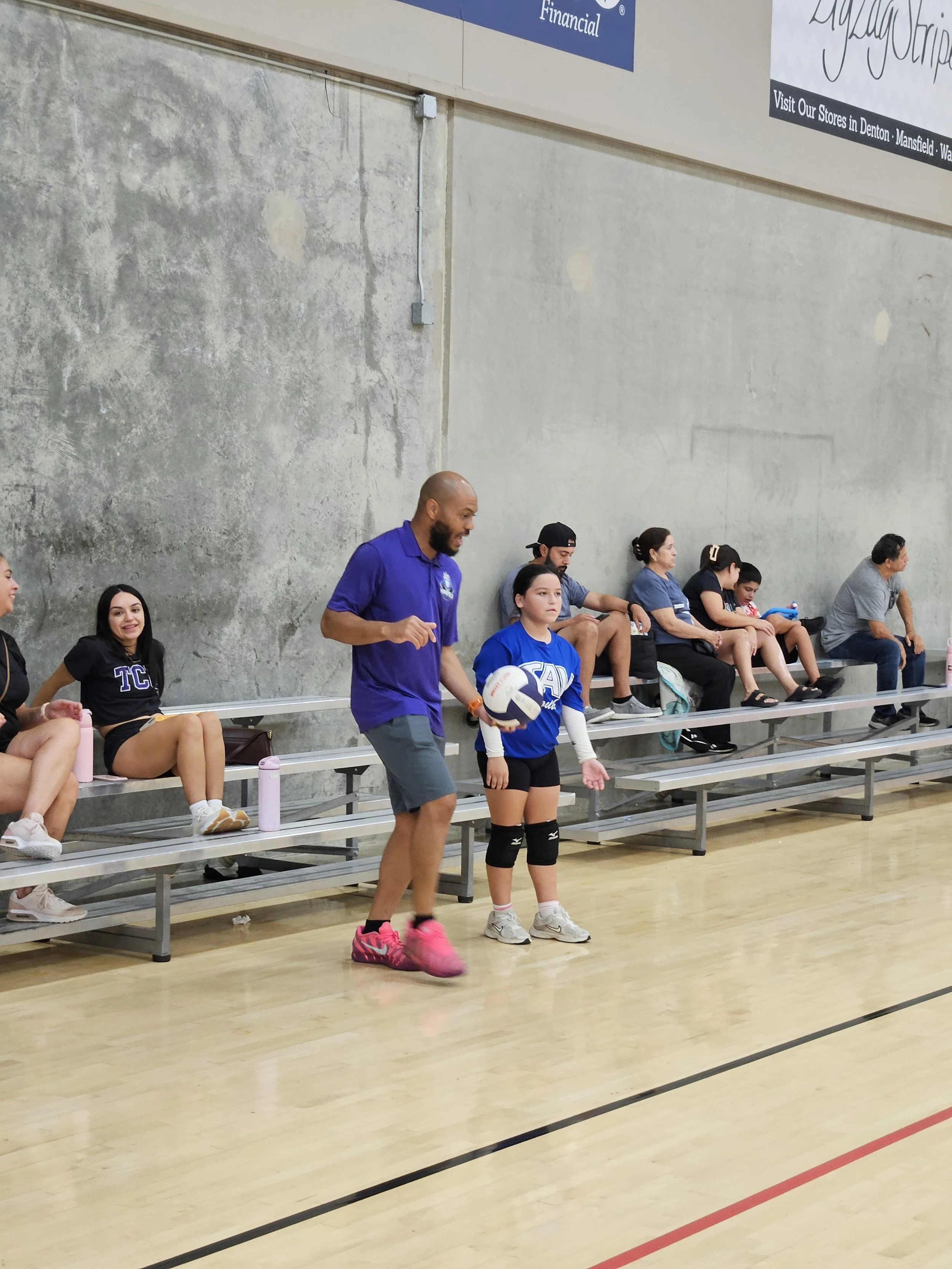 People seated on bleachers watching a volleyball game in a gym gymnasium, with one woman and one man standing and holding a volleyball.