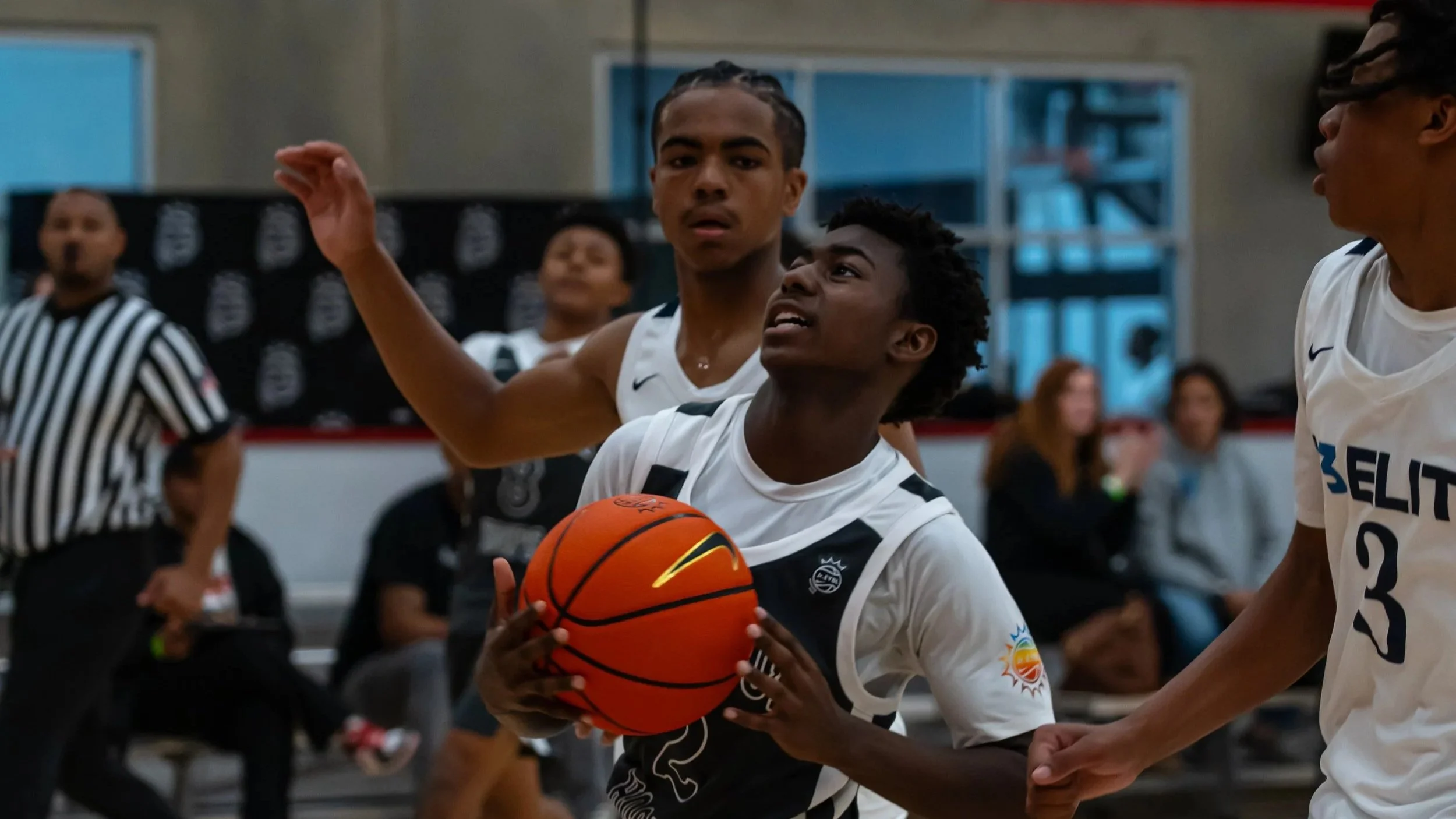 Two basketball players in white jerseys contest for possession of the basketball during a game, with an official and spectators in the background.