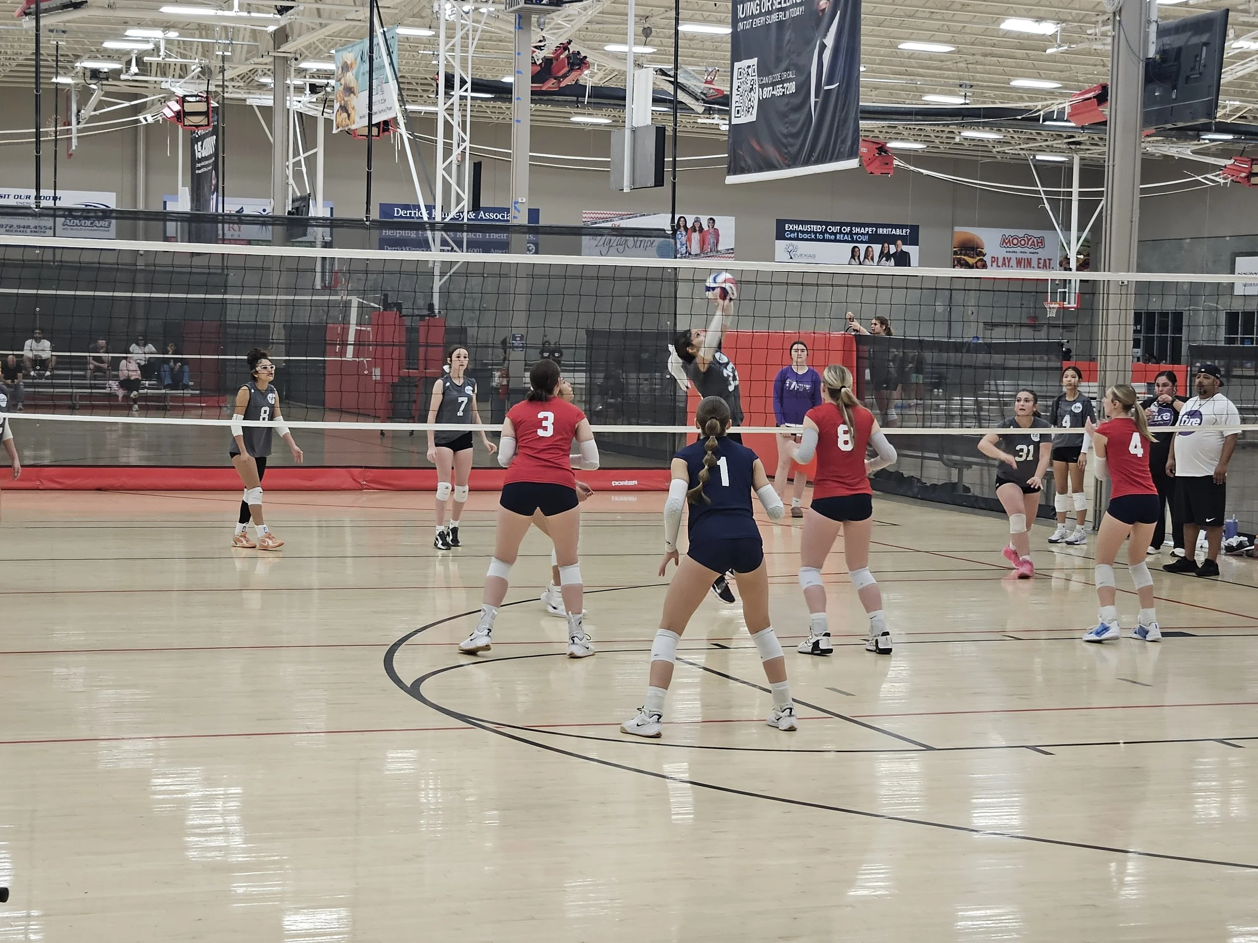 An indoor volleyball game is in progress with players on both teams preparing for a serve. The gym has a high ceiling, and there are banners and advertisements around. Several players are wearing jerseys and knee pads, and a net divides the court.