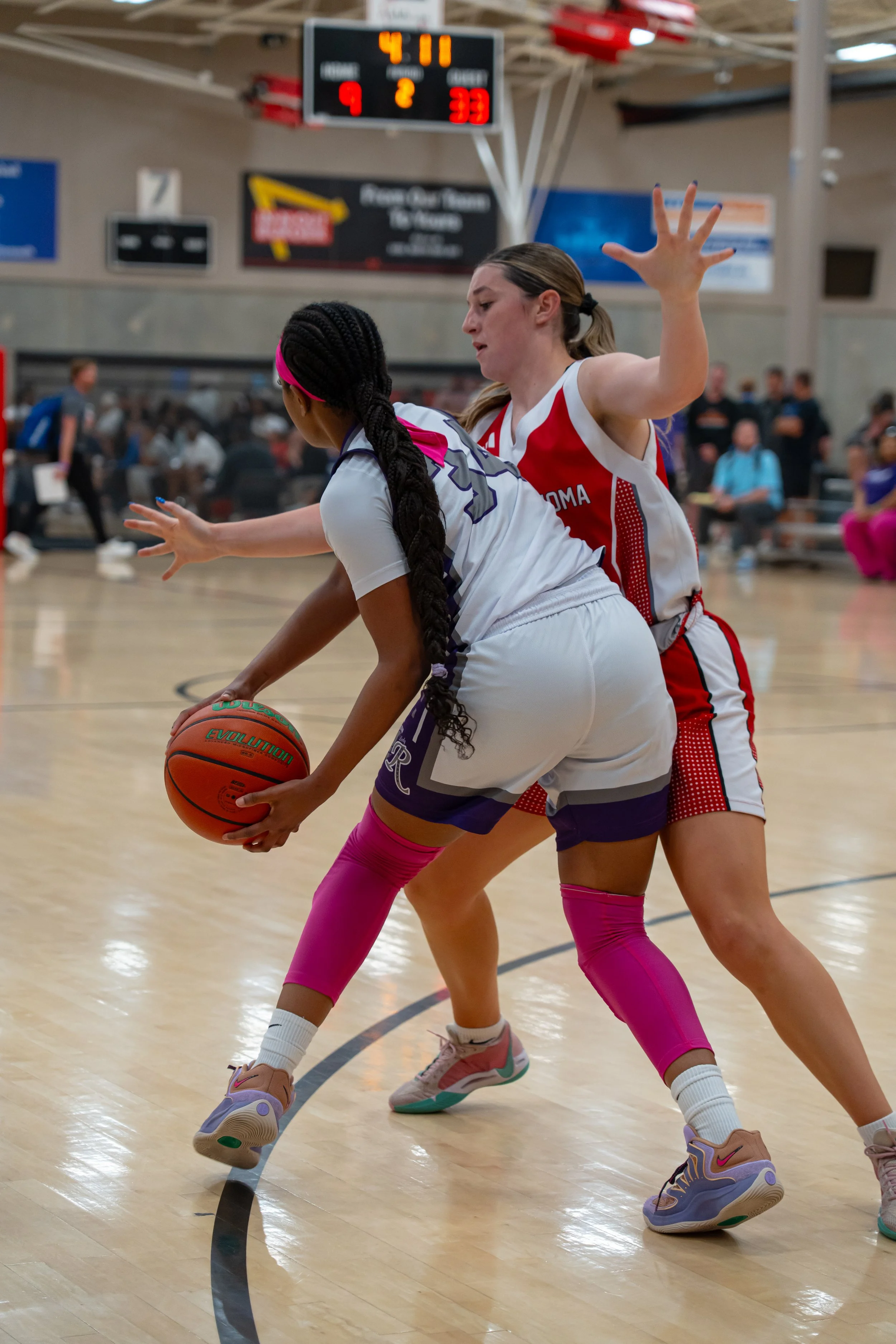 Two female basketball players in colorful uniforms battle for possession of the ball on an indoor court during a game, with spectators and a scoreboard in the background.