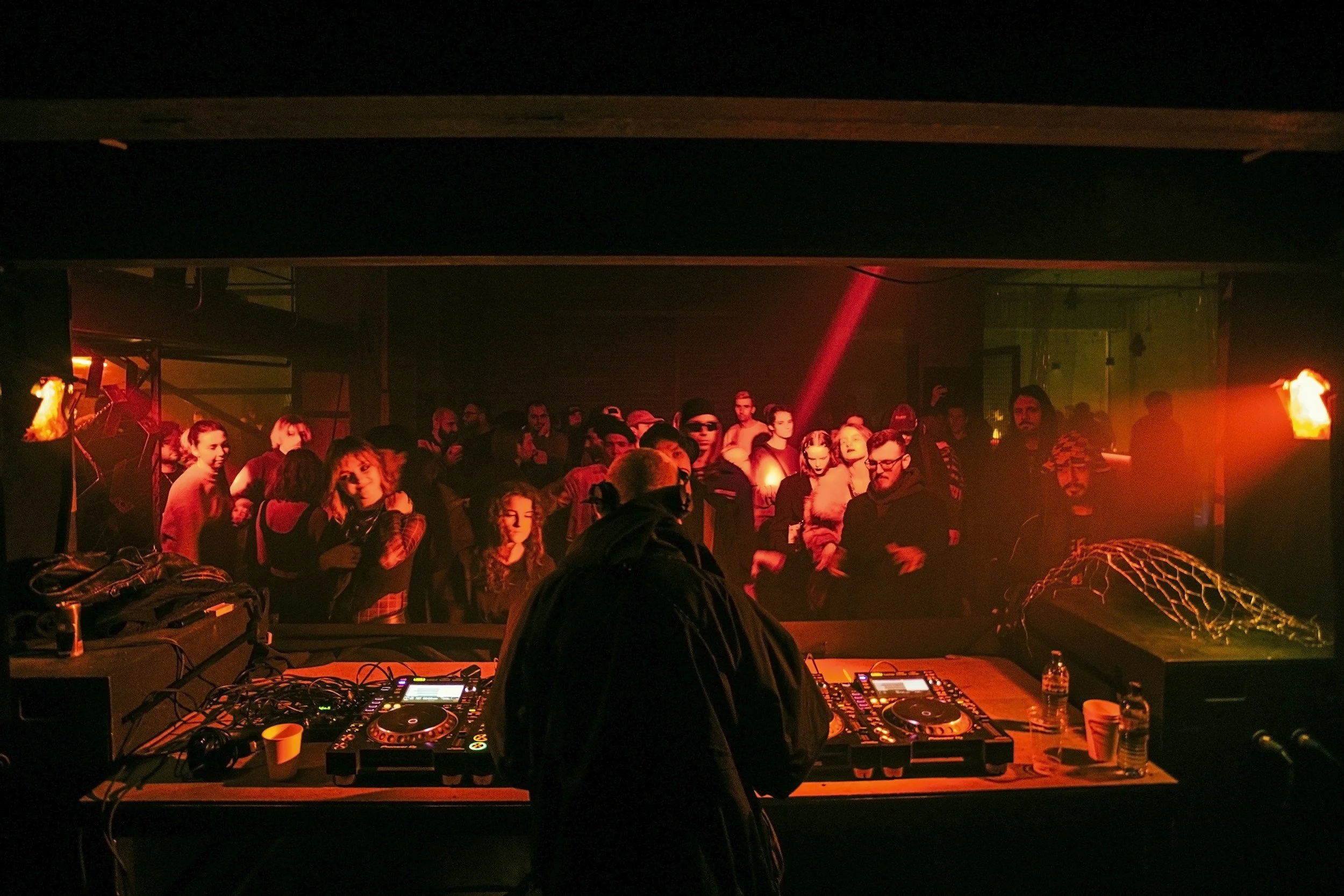 A DJ setup with turntables and mixers in the foreground. Behind, a crowd of people is gathered in a dimly lit room with red and yellow lighting, likely at a nightclub or concert event.